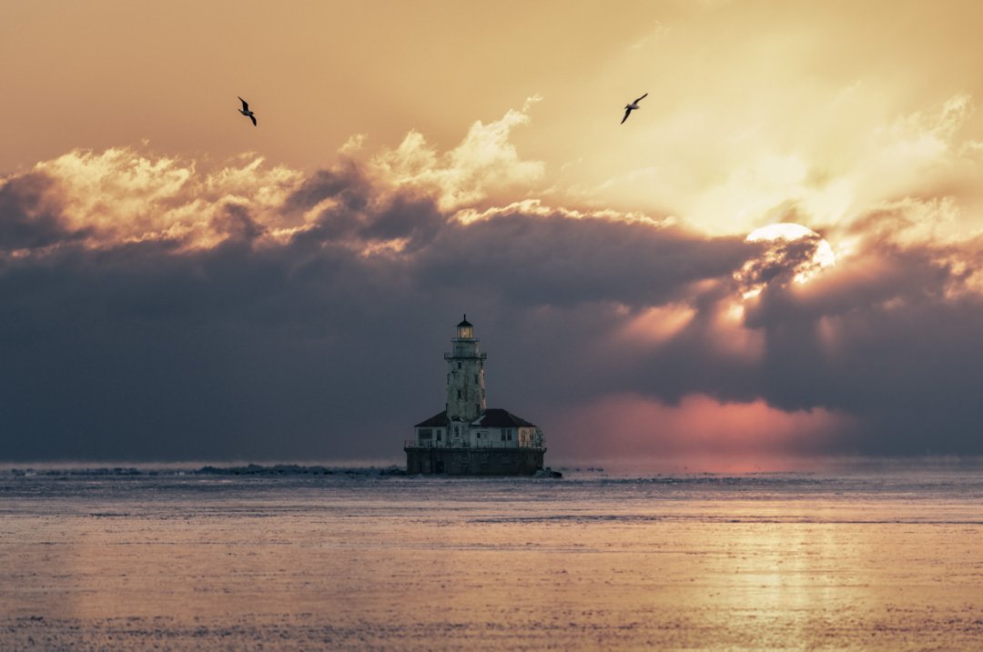 A serene sunset over a lighthouse with rays piercing through clouds and birds soaring in the sky.