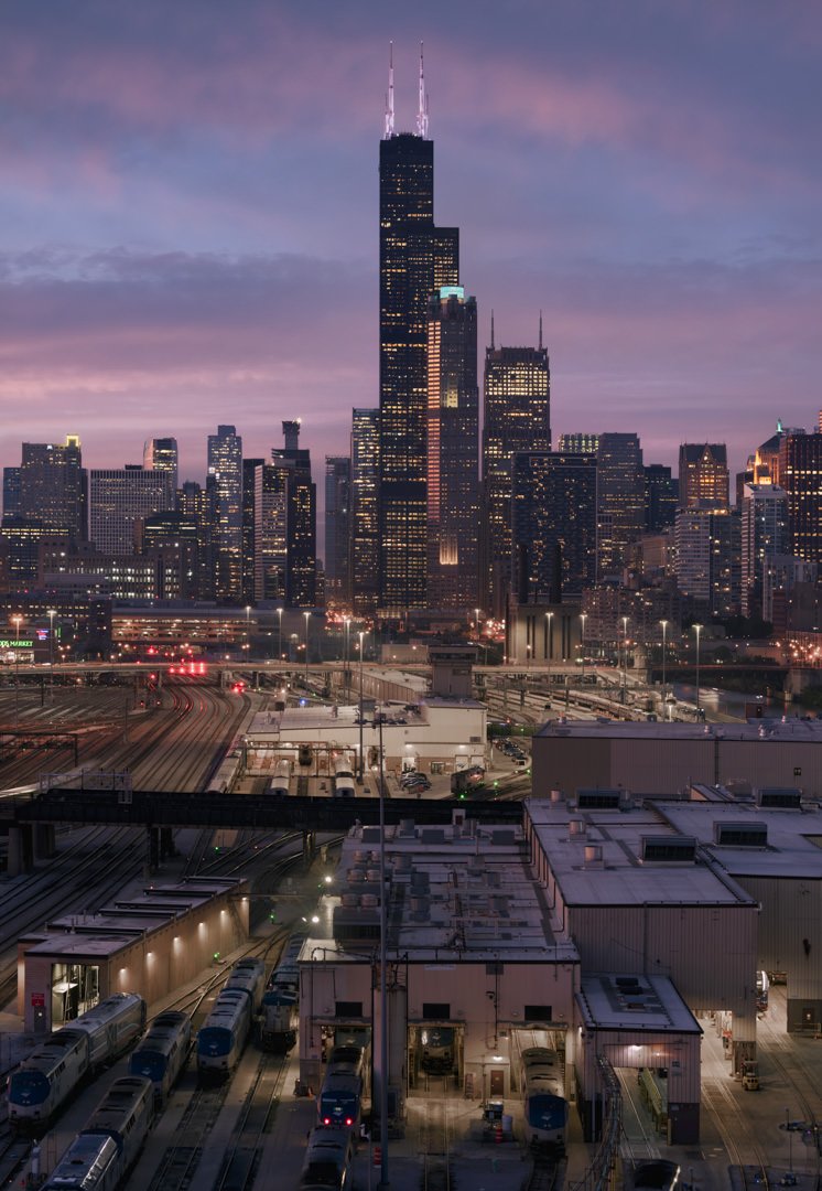 Dusk view of the Chicago skyline with Willis Tower prominent against a pastel sky, foregrounded by train tracks and industrial buildings.