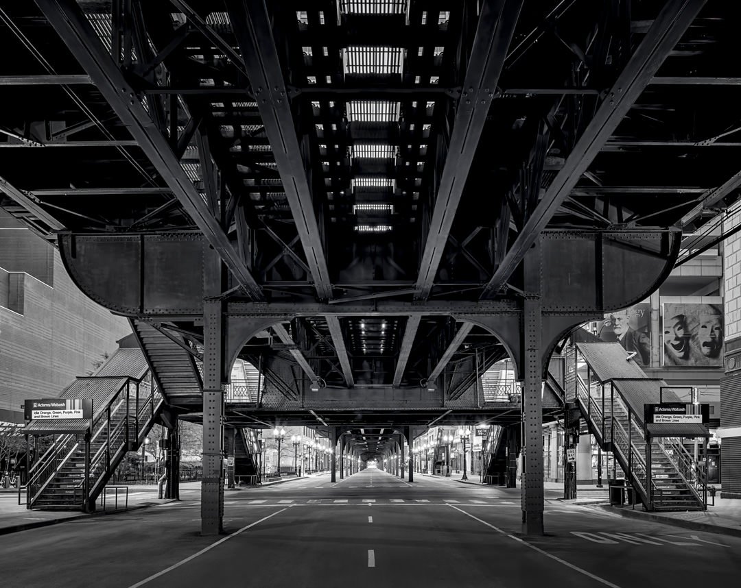 Elevated train tracks in grayscale over an empty Chicago street with staircases on either side and city lights casting shadows.