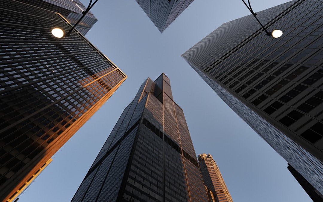 Looking up at a towering cluster of skyscrapers against a clear blue sky, with the setting sun casting a warm glow on the facades, and a single street lamp glowing in the foreground.