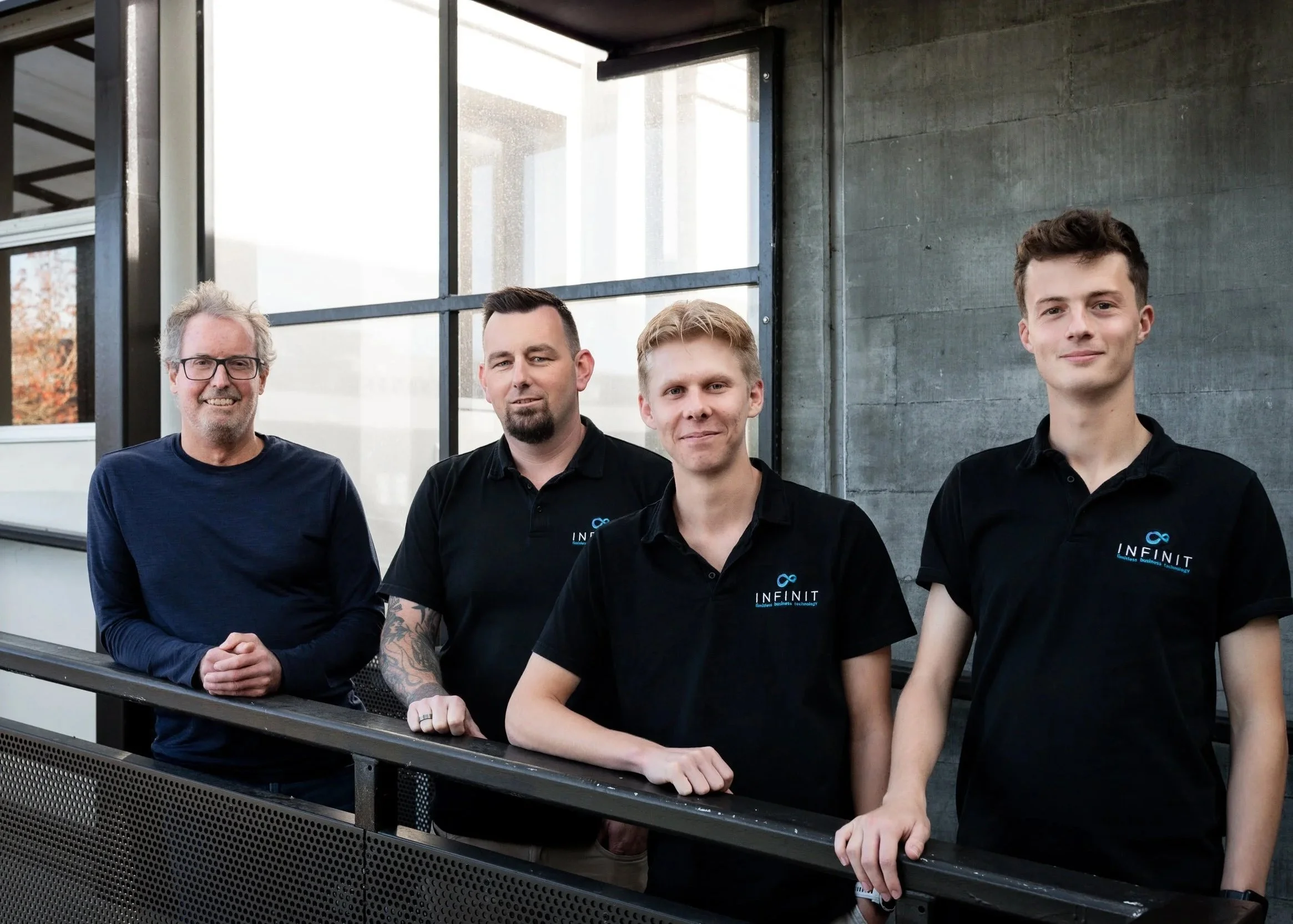 Four men standing on a balcony or terrace outside a modern building, smiling and looking at the camera. Some wear black shirts with a logo, and one wears glasses.