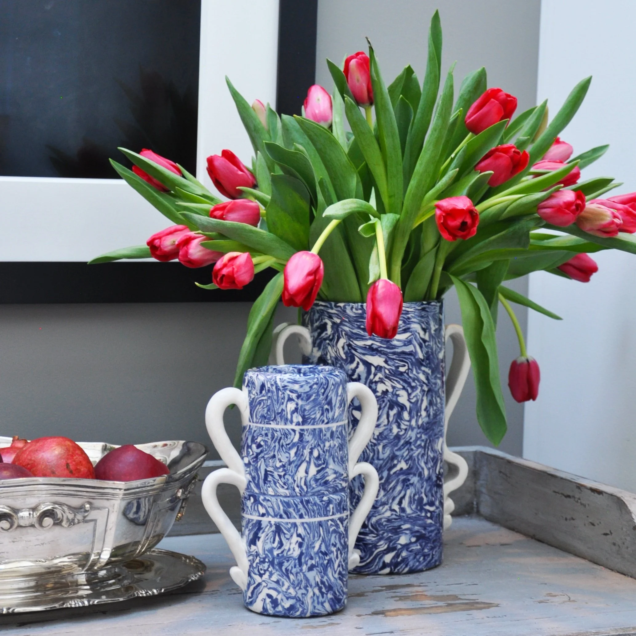 Red tulips in a blue and white marbled vase beside a stack of matching cups and a silver fruit bowl on a wooden surface.