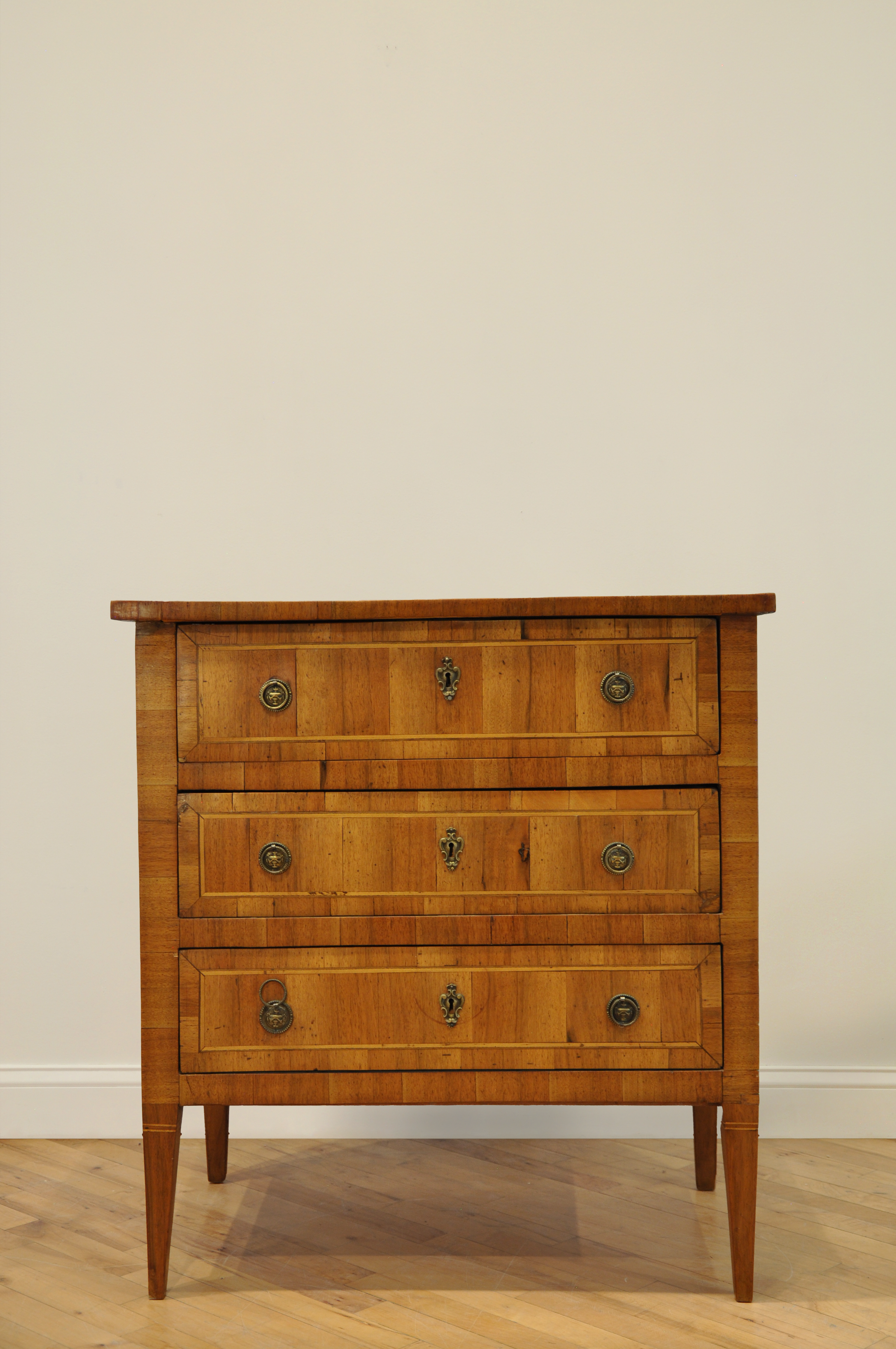 Wooden chest of drawers with three drawers and brass handles, set against a plain wall on a wooden floor.