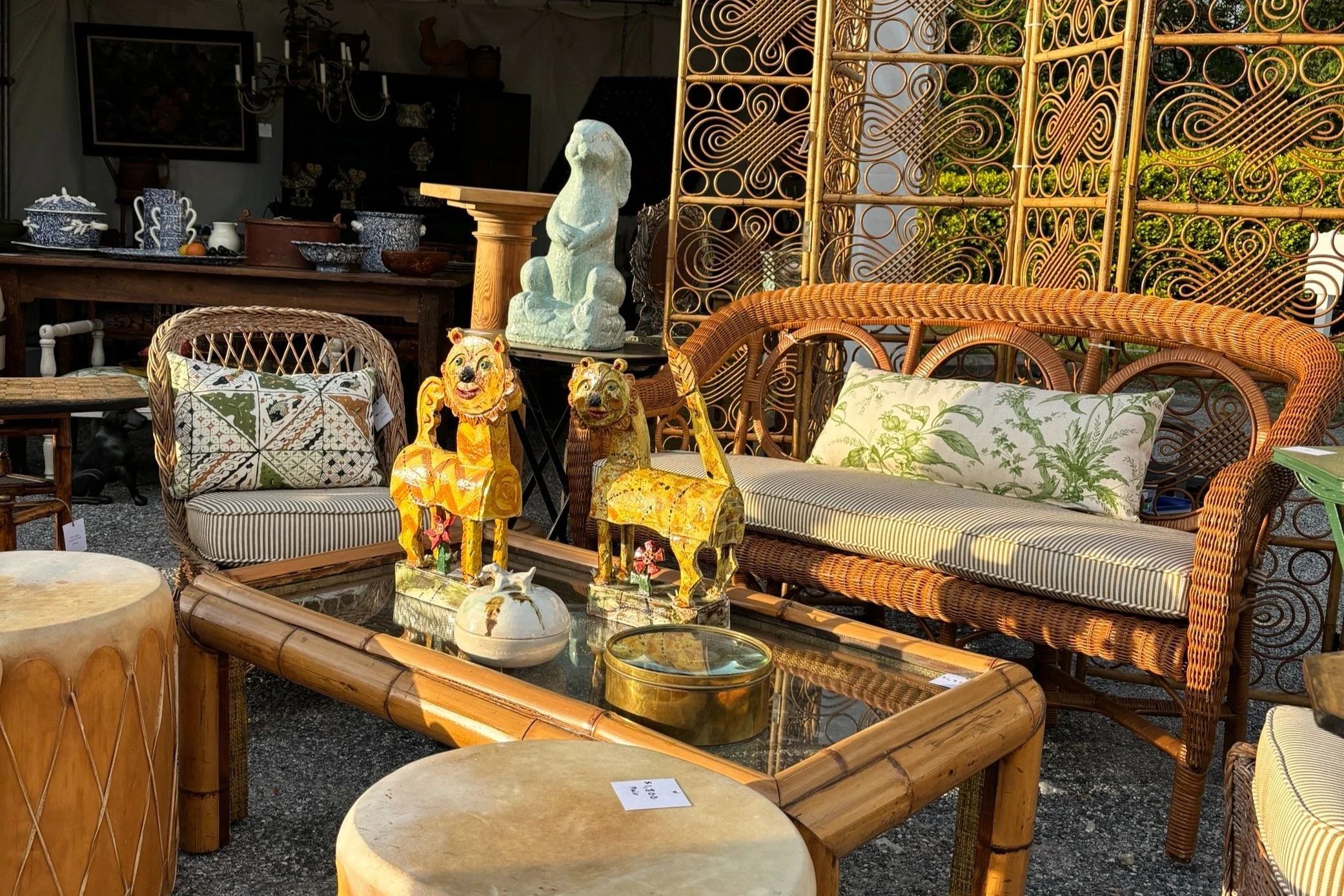 Outdoor antique market with wicker furniture, decorative lion sculptures, and various pottery pieces. Bamboo coffee table with glass top and cushions on a wicker sofa and chair.
