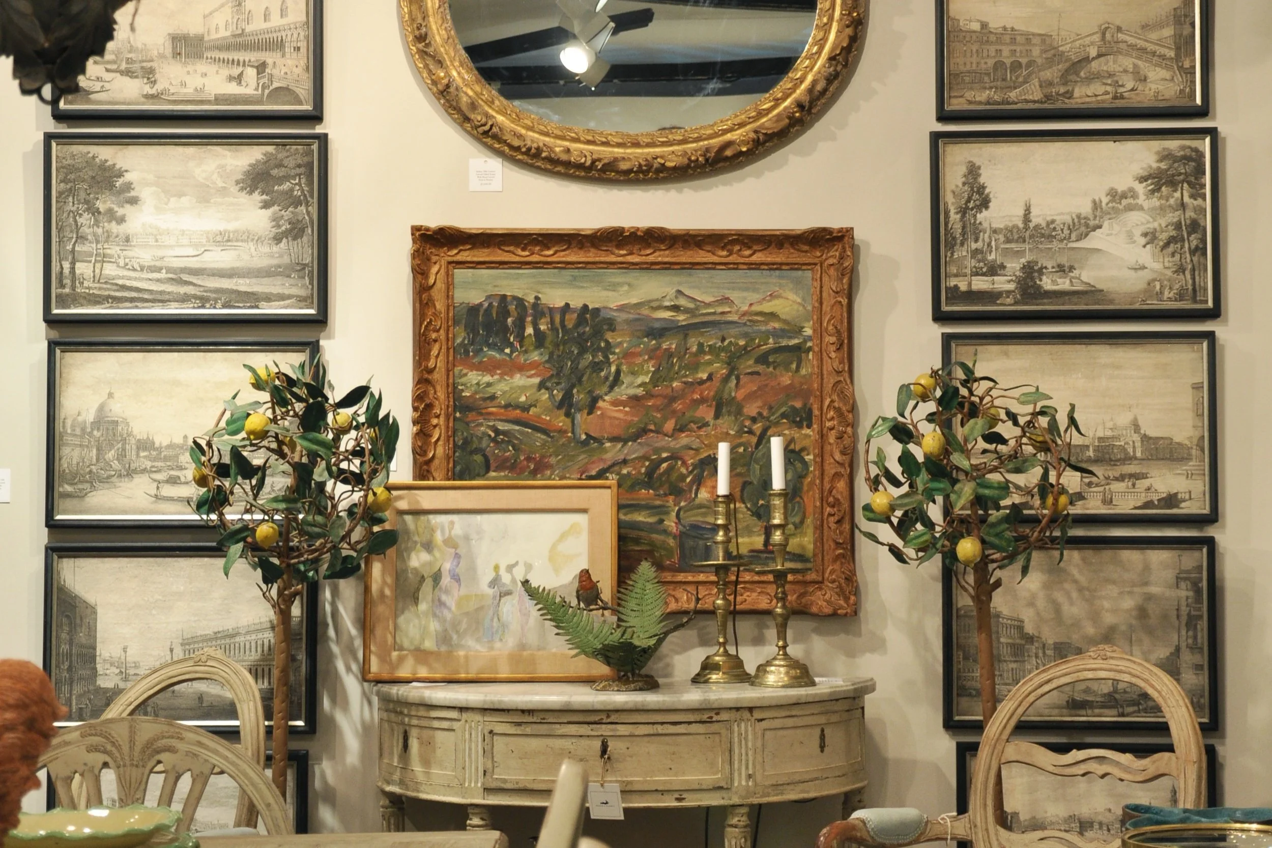 Wall art and decor in a vintage interior setting, featuring various framed paintings and sketches, a round ornate gold mirror, a small decorative table with candles, a decorative plant, and a fern in a vase.
