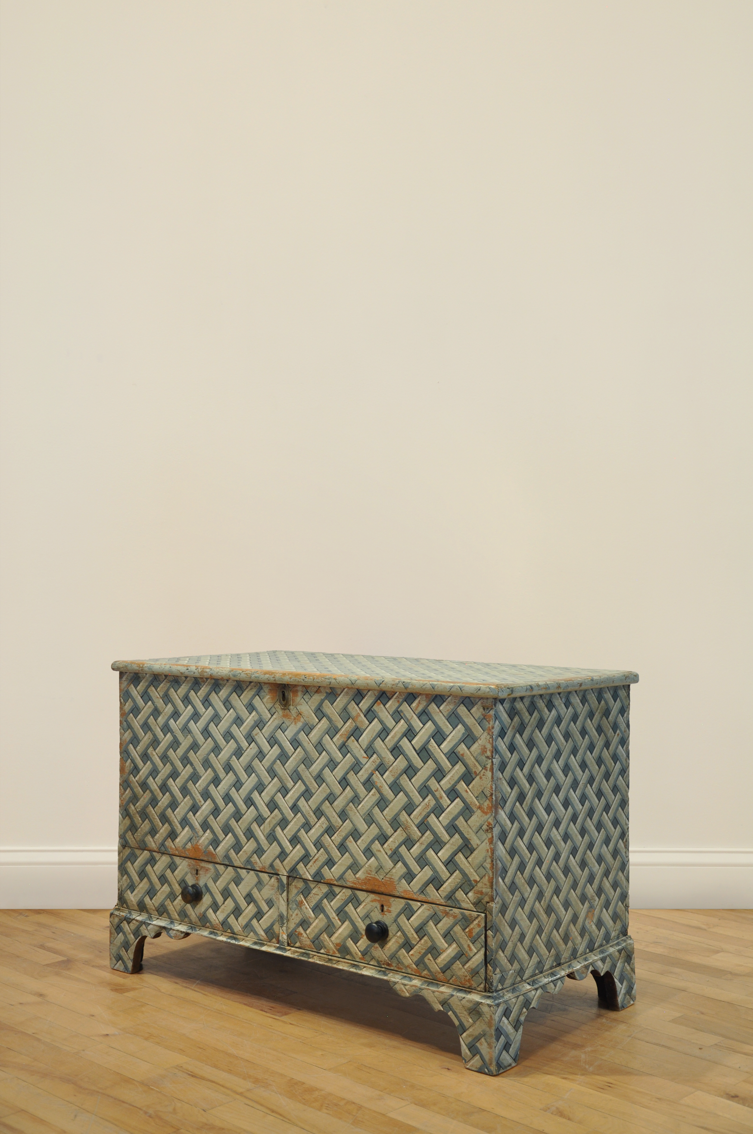 Blue woven pattern wooden chest against a beige wall on a wood floor.