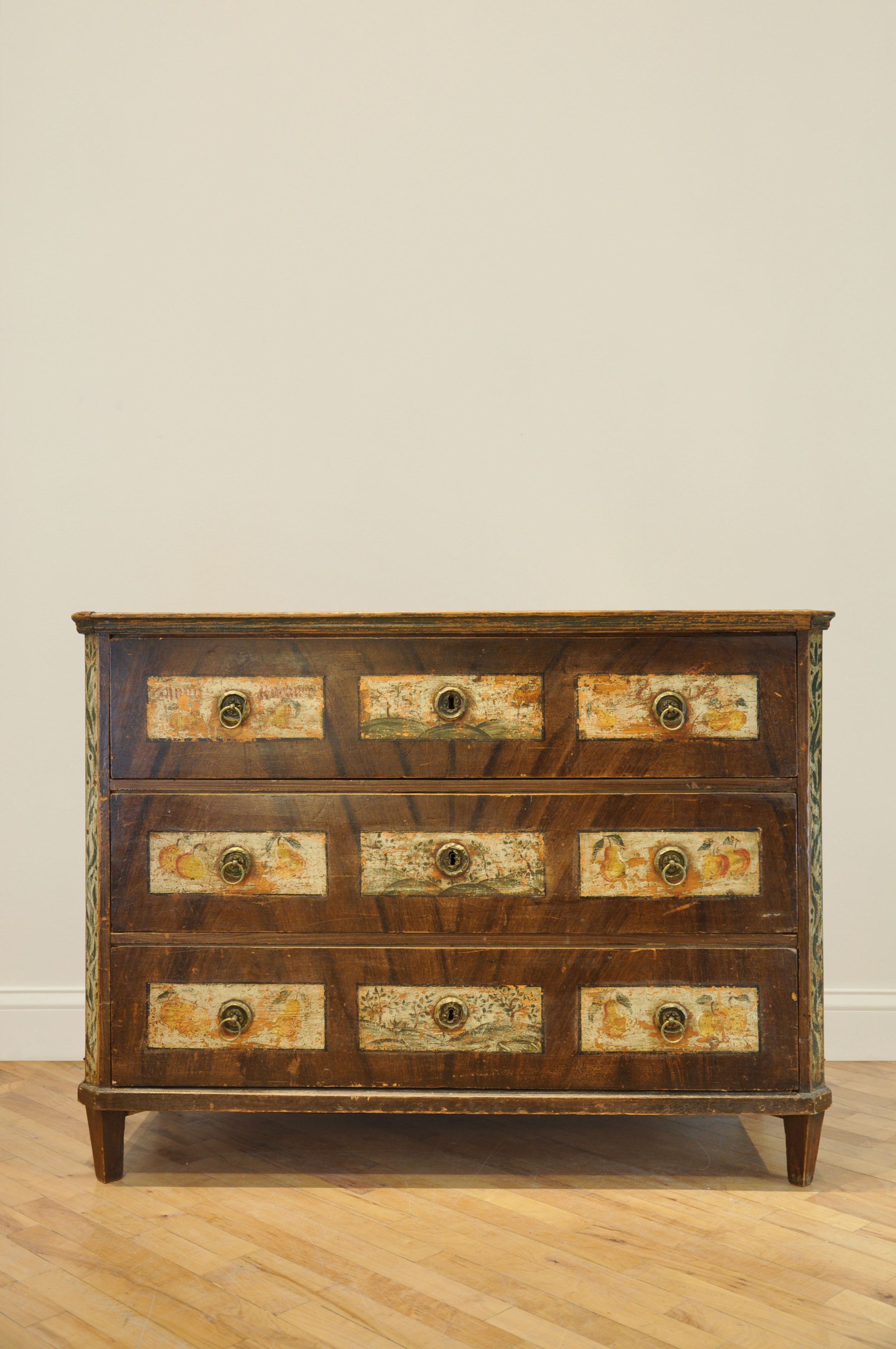 Antique wooden dresser with three drawers featuring vintage painted designs, brass handles, and a parquet floor.