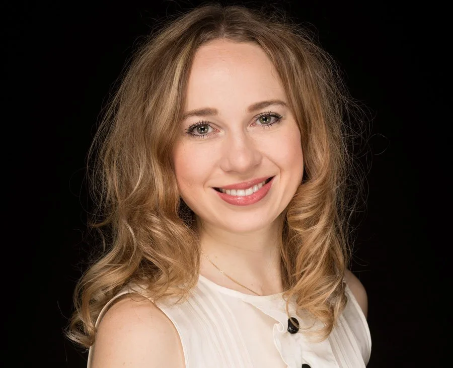 Portrait einer lächelnden jungen Frau mit blonden, lockigen Haaren vor schwarzem Hintergrund.