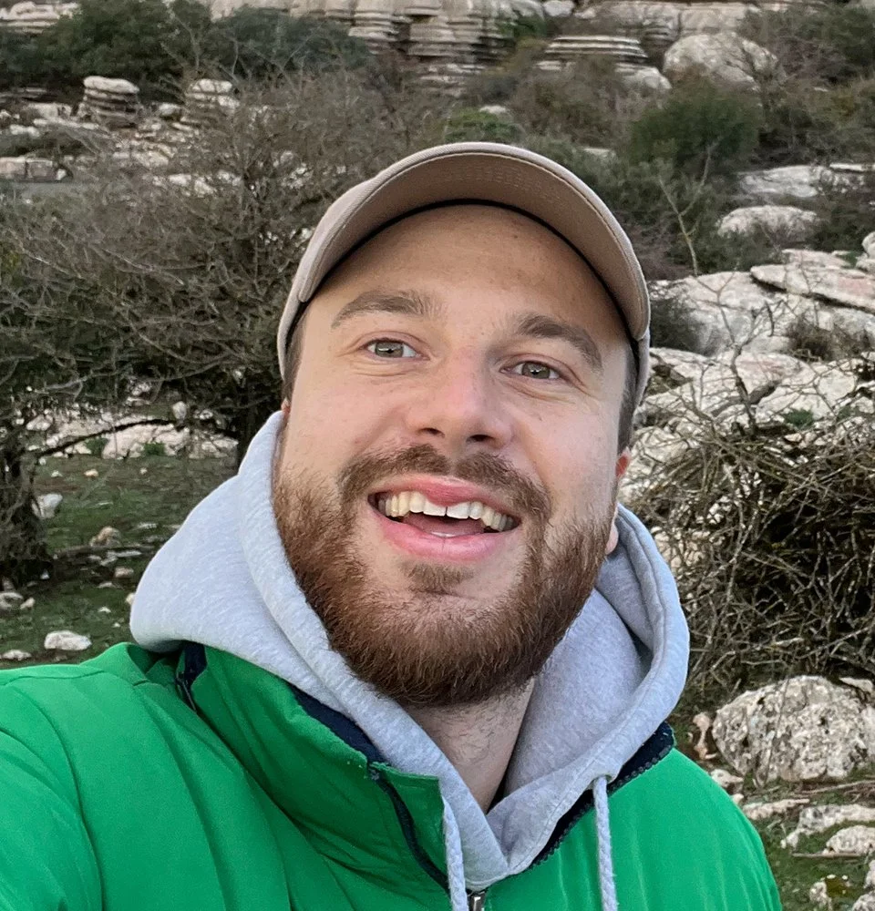 Ein lachender Mann mit Bart, trägt einen grauen Hoodie, eine grüne Jacke und eine beige Kappe, steht vor einer natürlichen, felsigen Landschaft mit Büschen im Hintergrund.