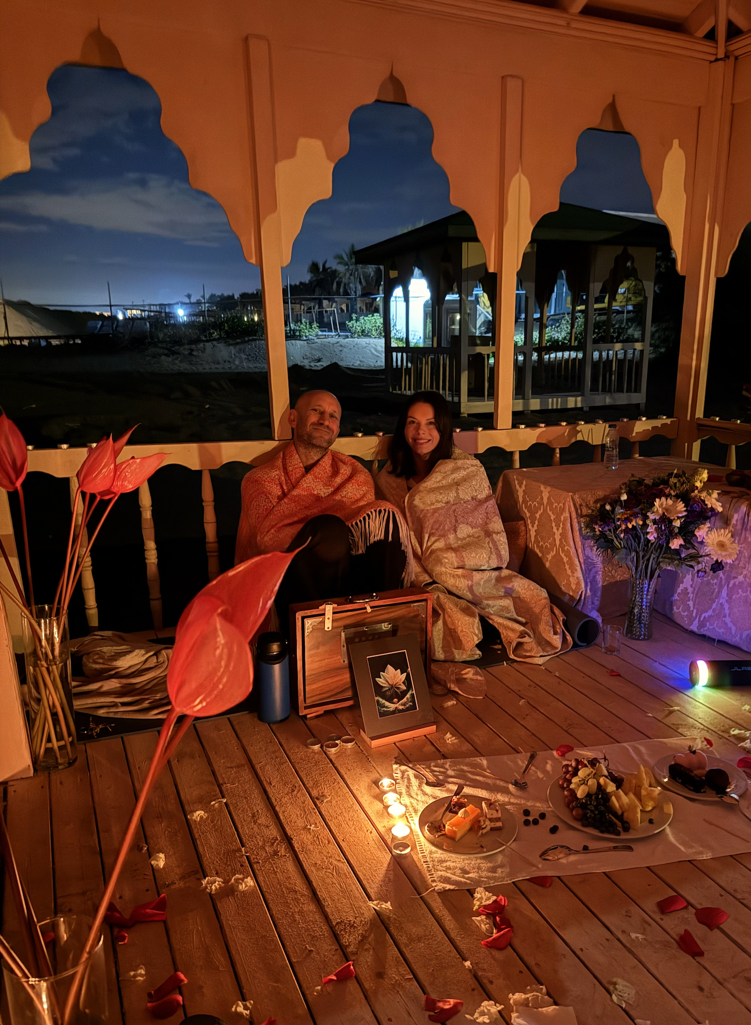 Two people sitting on a wooden floor inside a decorated tent at night, with flowers, candles, and food around them, and outdoor scenery visible through the open sides of the tent.