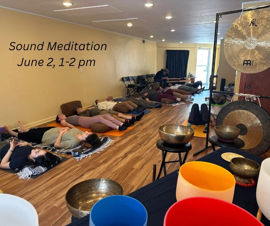 A group of people lying on mats and cushions in a room, participating in a sound meditation session. The room has wooden floors, a yellow wall, and a variety of singing bowls and gongs in the foreground. Text on the wall reads 'Sound Meditation June 2, 1-2 pm'.