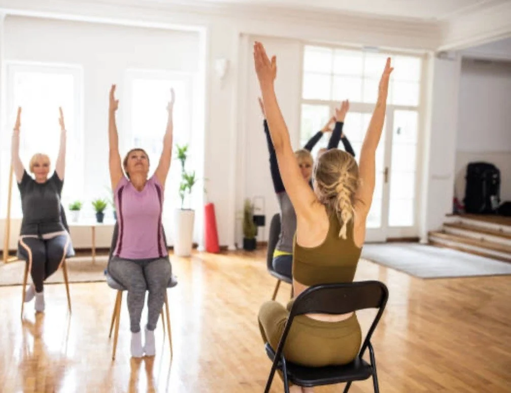 Group of women participating in a seated yoga or meditation class in a bright, spacious room with wooden floors and large windows.