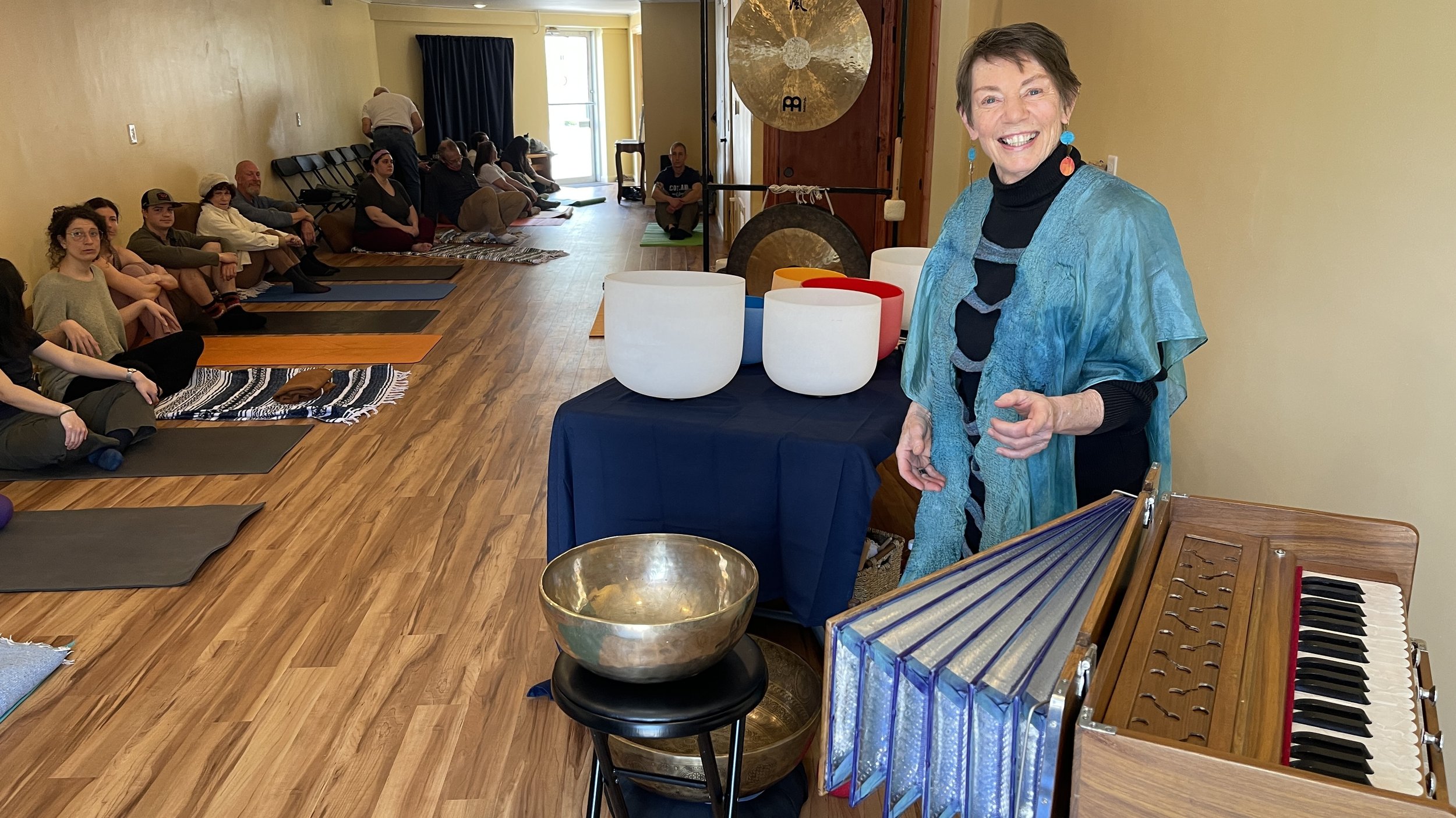A smiling woman standing next to a table with singing bowls and a small harmonium, with a group of people sitting on yoga mats in a room.