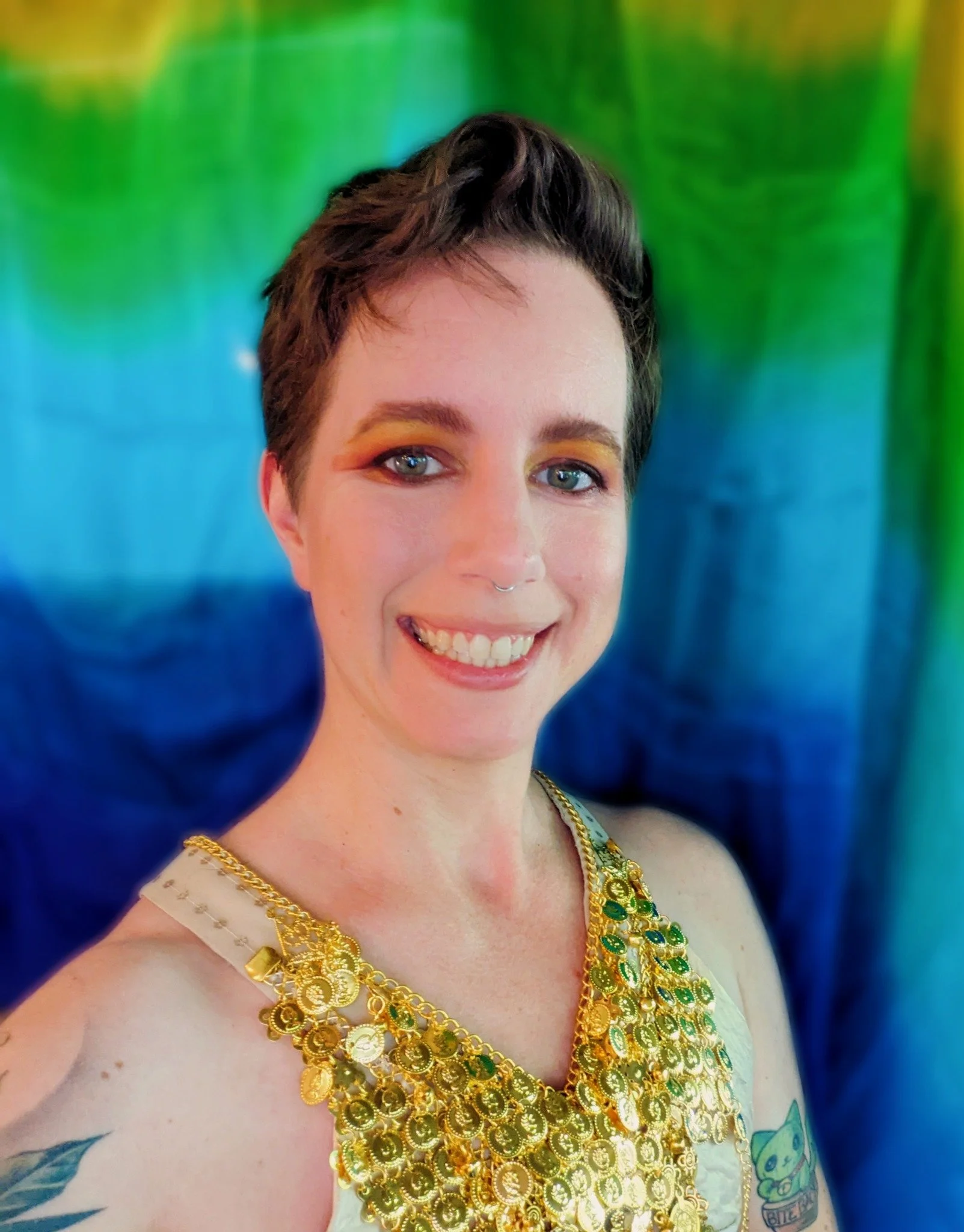 Woman with short dark hair and bright makeup, smiling, wearing a large gold necklace and a sleeveless top, standing in front of a rainbow-colored backdrop.