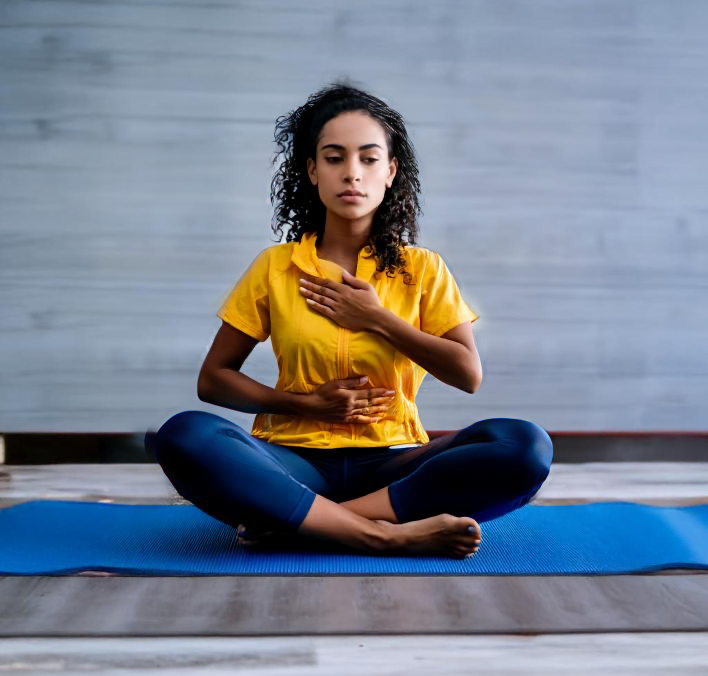 A woman practicing yoga indoors, sitting cross-legged on a blue mat with hands on her chest and stomach, wearing a yellow top and dark leggings.
