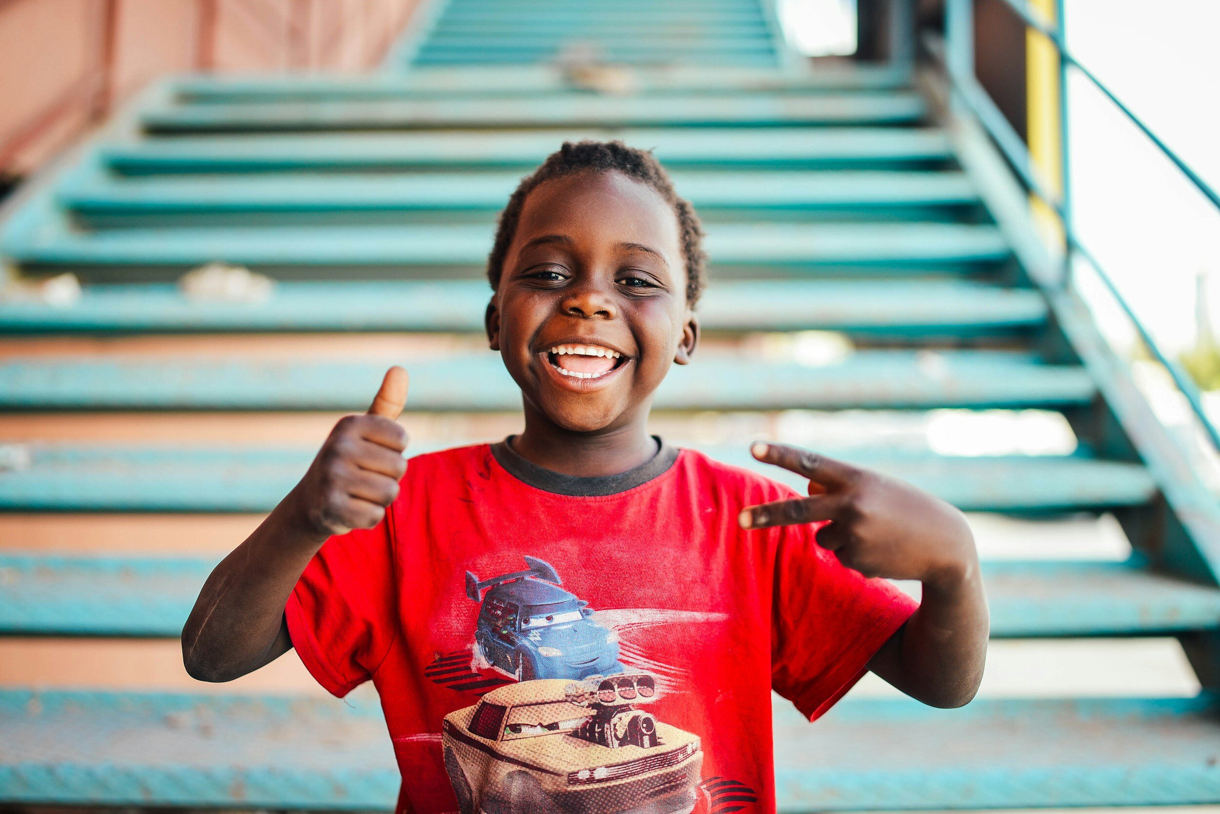 A happy young boy with short curly hair smiling and making a thumbs-up and peace sign with his hands, standing in front of a teal staircase.
