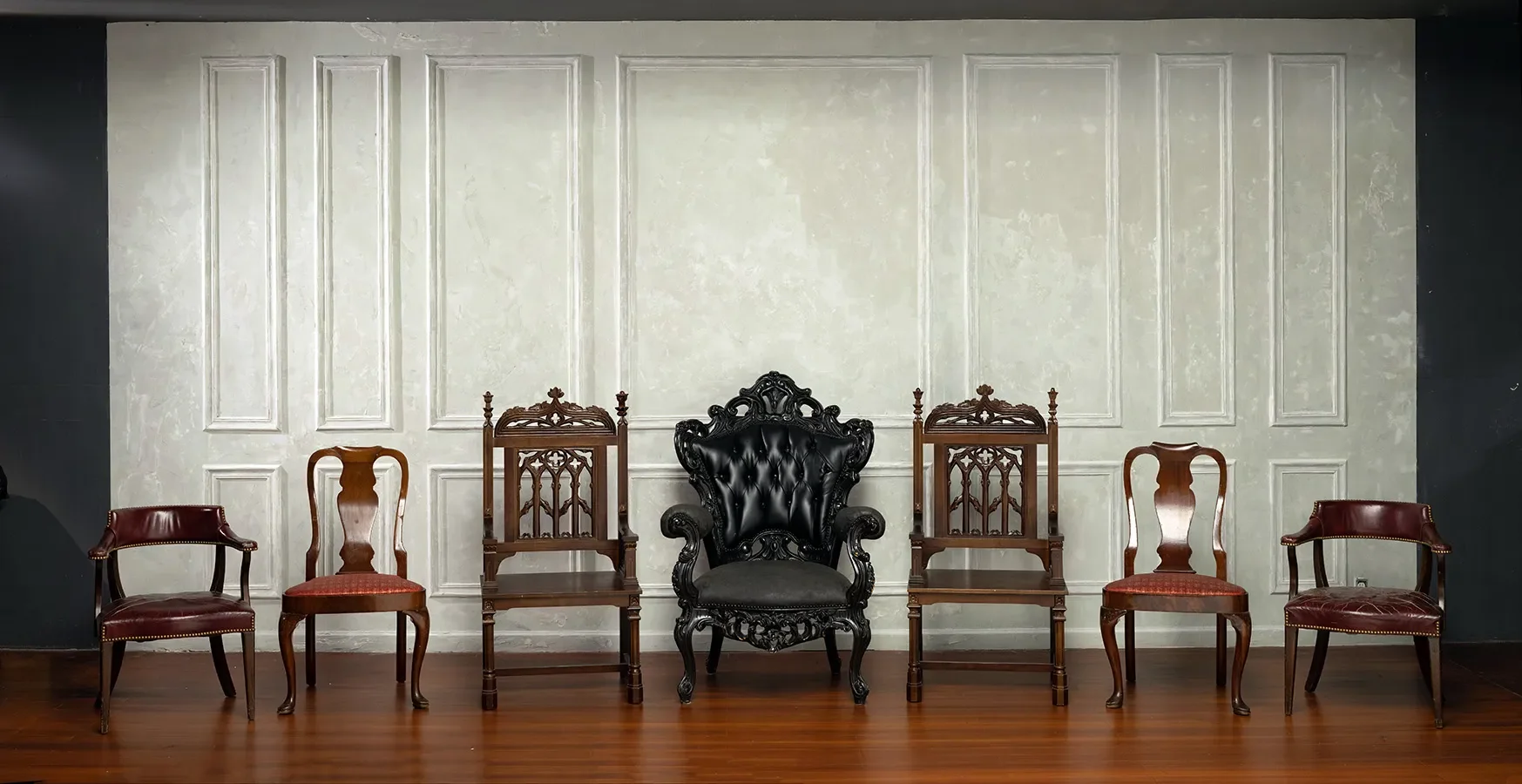 A row of antique and vintage chairs arranged against an ornate off-white paneled wall, with a large black tufted throne chair centered among the different wooden chairs.