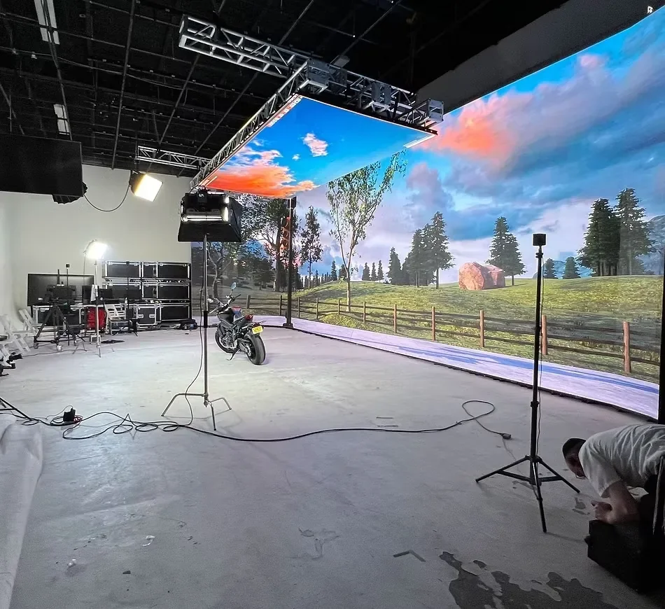 Virtual production studio featuring a curved LED wall, overhead lighting grid, and motorcycle set against a scenic digital landscape.