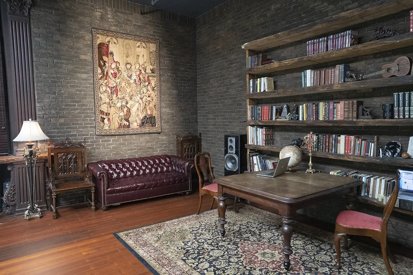 A cozy vintage study set with a wooden desk, burgundy leather sofa, rustic bookshelves, a decorative tapestry, and assorted books and antique decor against dark brick walls.