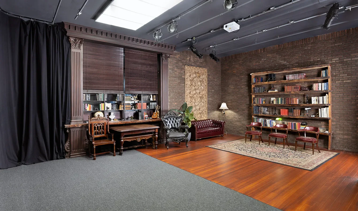 A wide view of a studio set combining a classic study area with brick walls, wood floors, a large library backdrop, antique furniture, and overhead production lighting.