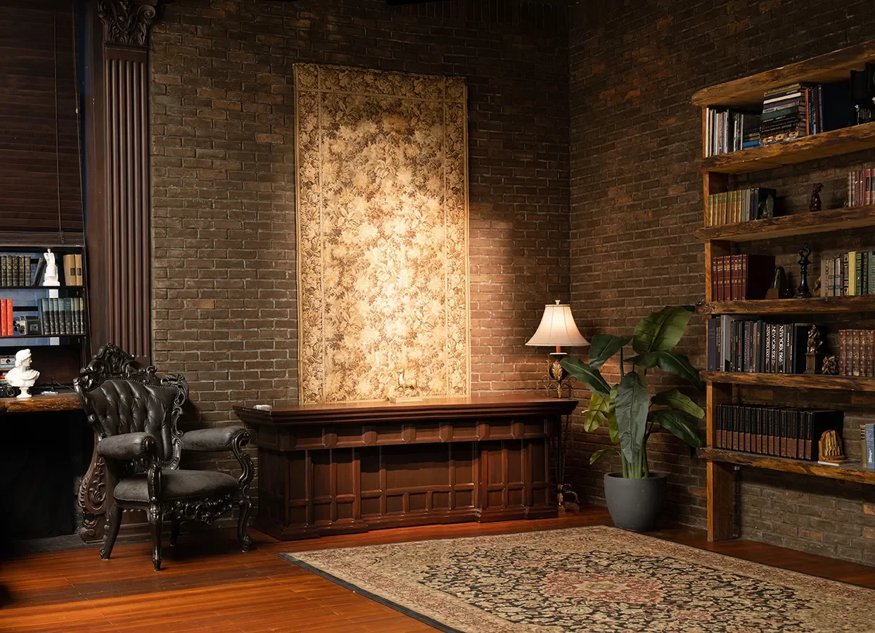A vintage-style study set with dark brick walls, a carved wood desk, tufted leather chair, tall bookshelves filled with books, warm lamps, and a large decorative tapestry hanging on the wall.