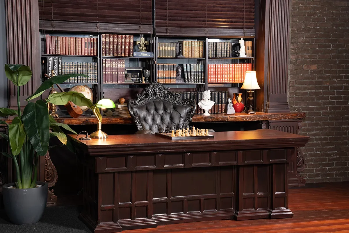 A classic study set featuring a large carved wooden desk, an ornate tufted leather chair, tall bookcases filled with books and vintage objects, and warm lighting from desk and table lamps.