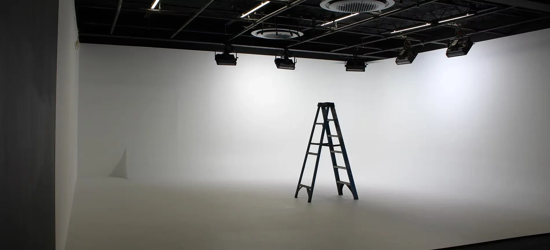Spacious white cyclorama studio with seamless curved walls, overhead lighting grid, suspended studio lights, and a ladder for production setup.