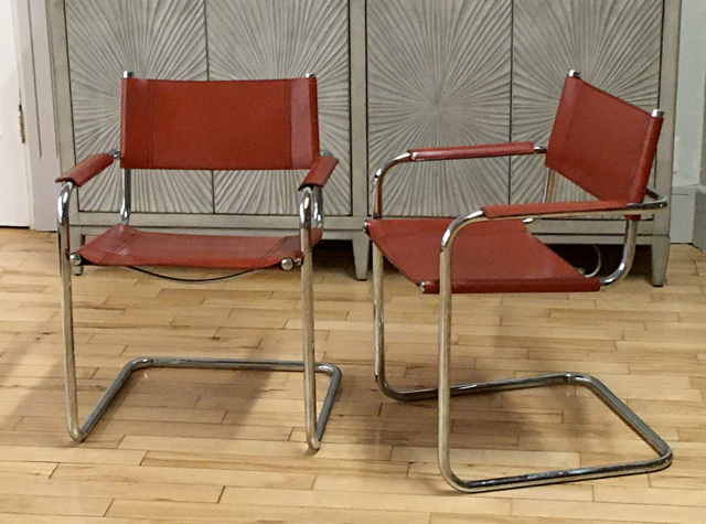 Two vintage modernist side chairs with chrome frames and red leather upholstery, positioned on a wooden floor in front of a decorative wall panel.