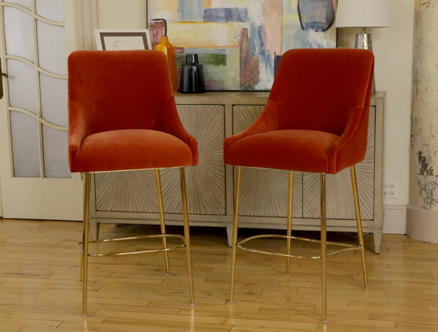 Two orange velvet bar stools with gold legs in front of a beige cabinet with colorful abstract artwork and decorative items, in a room with wooden flooring and a painted wall.