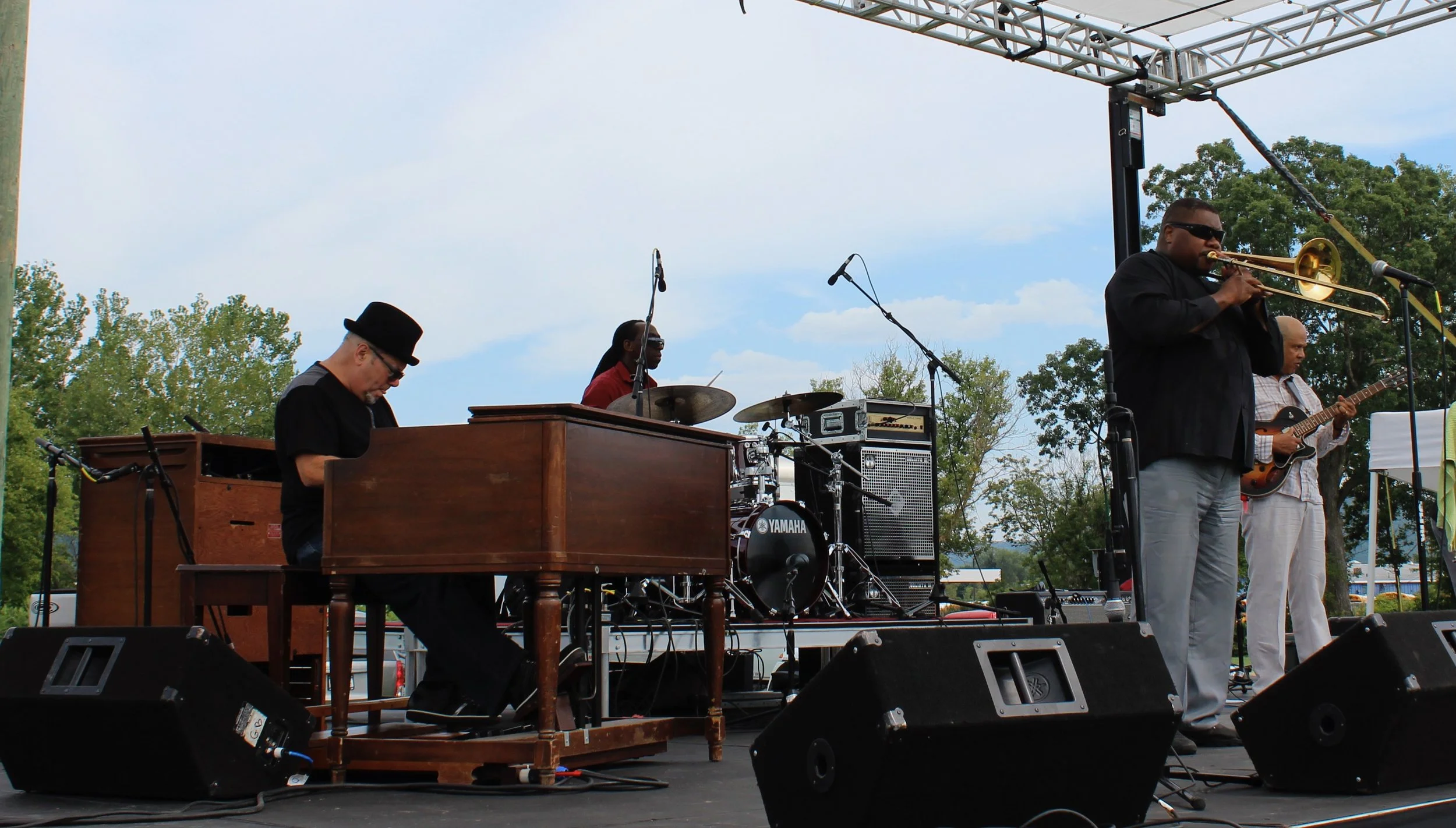 Live outdoor jazz band performance with an organ player, drummer, trombone player, and guitarist on stage.