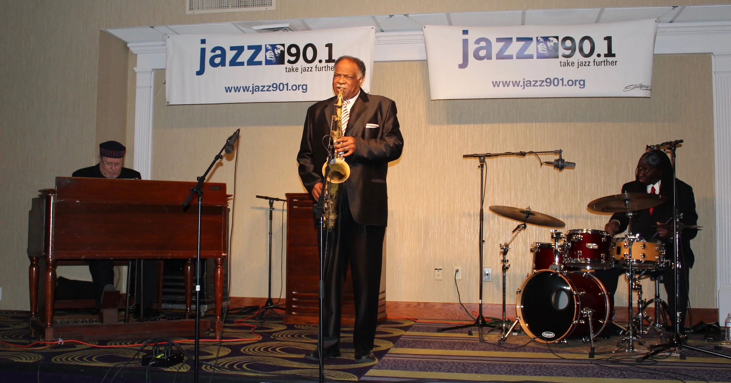 A jazz trio performing on stage. The saxophonist is standing in the center, flanked by a drummer on the right and an organist on the left. Banners above indicate the event is sponsored by Jazz 90.1."