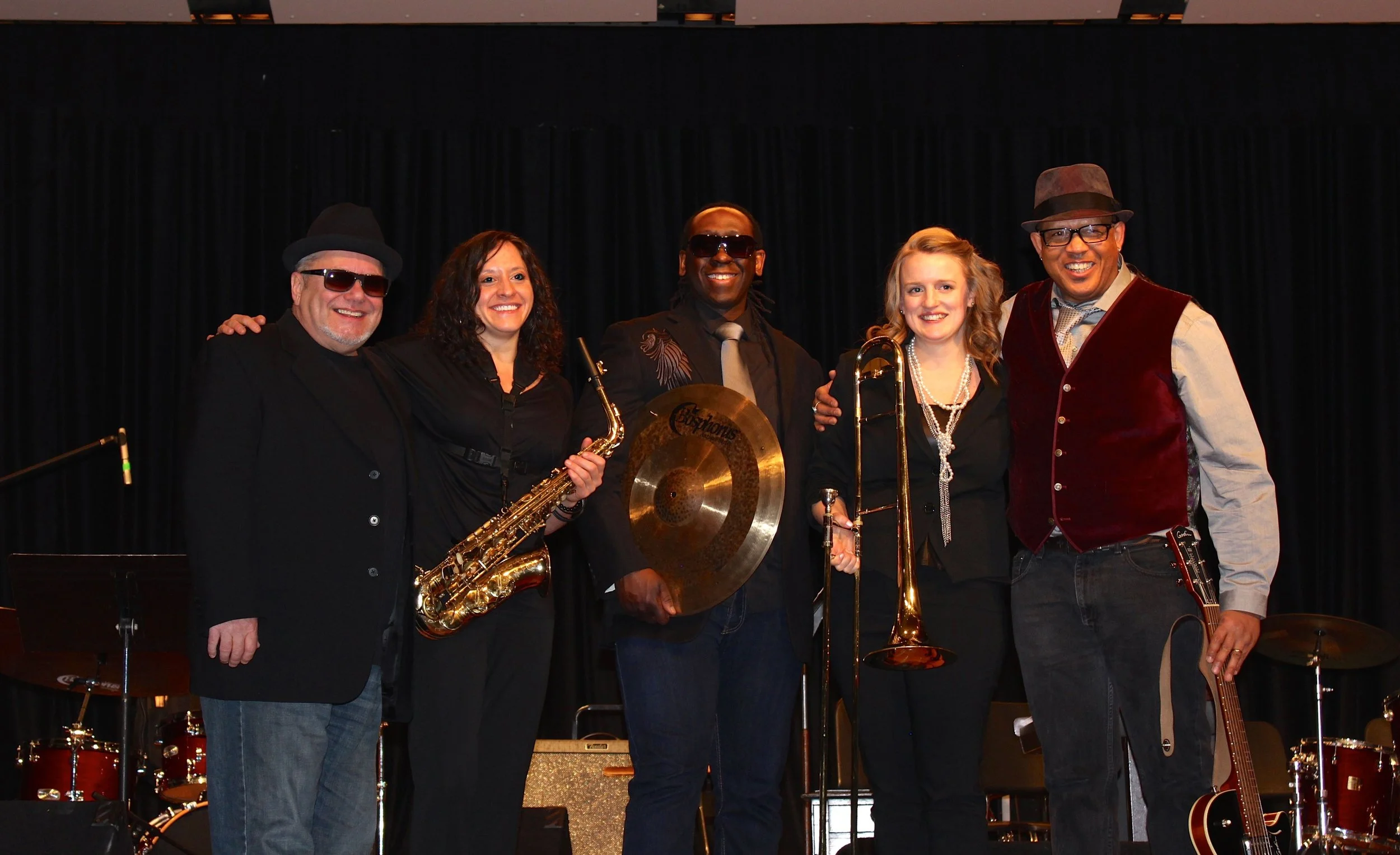 Group of five musicians on stage holding instruments including saxophone, trombone, and cymbal, with drums and guitar in the background.