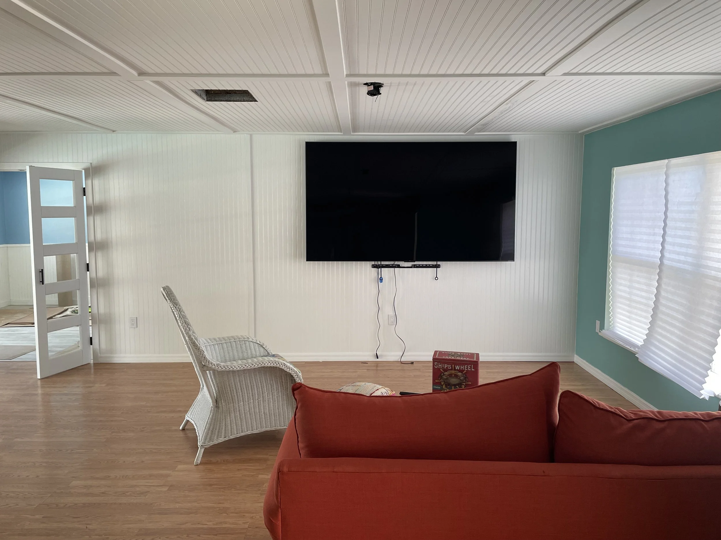 Living room with a wall-mounted flat-screen TV, white wicker chair, red couch, and wooden flooring. Light blue and white walls with blinds on windows, partially open, letting in natural light.