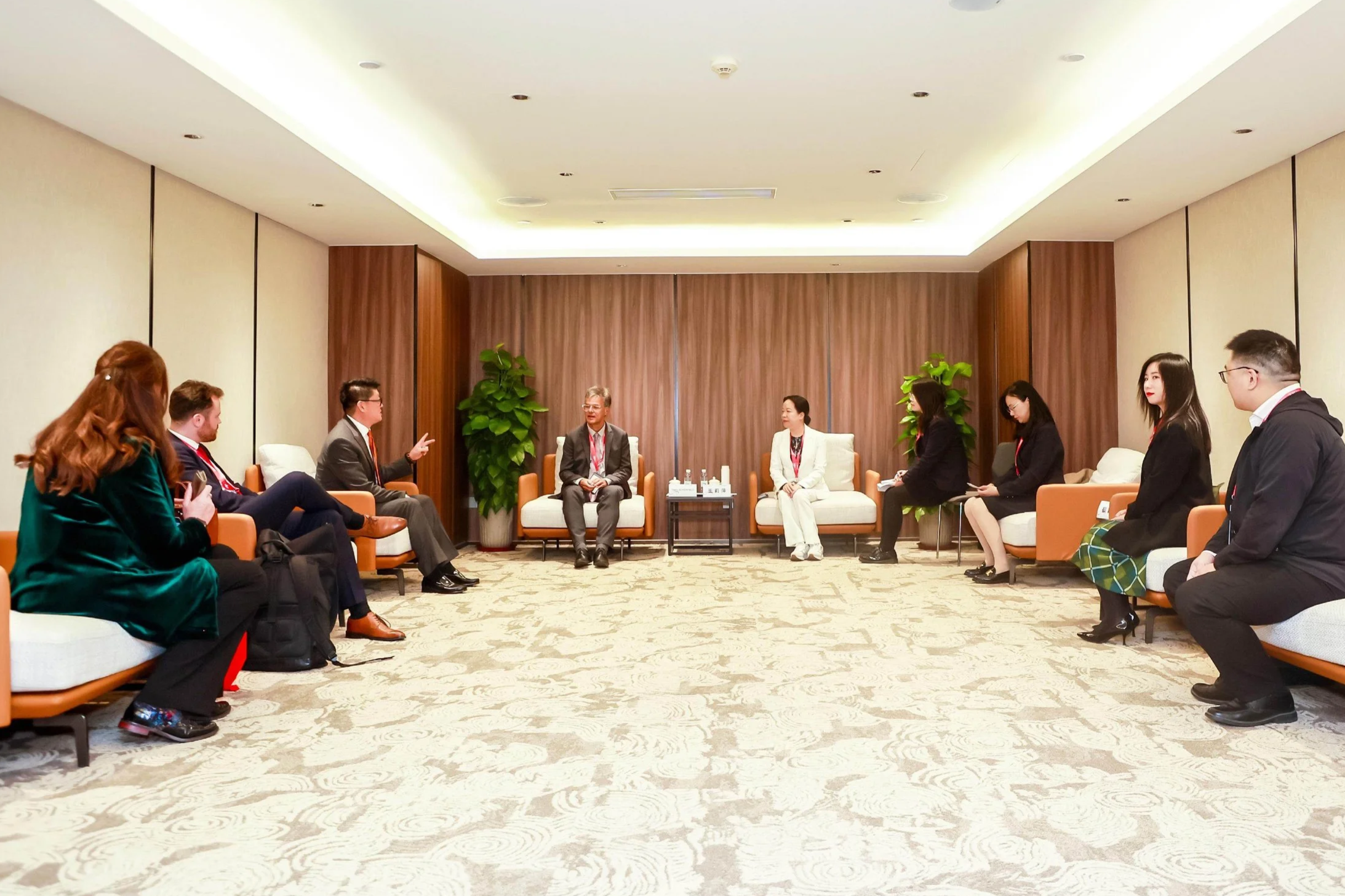 A business meeting in a modern conference room with eight people seated on sofas and chairs. The room has a wooden backdrop, green plants, and logos for IASC and Global Aerospace Cluster Partnership on the wall.