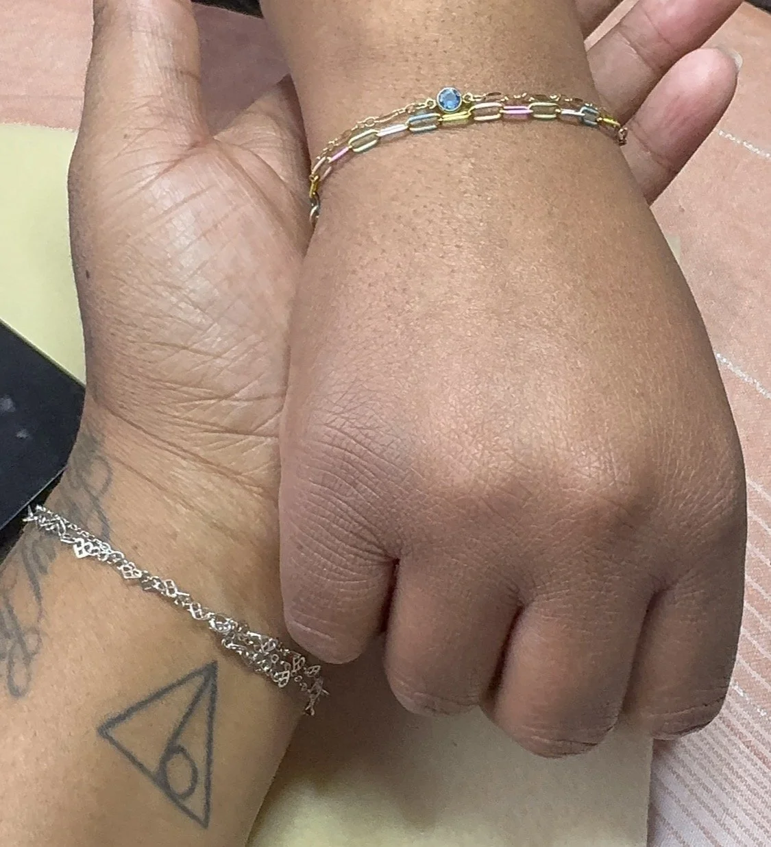 Close-up of two intertwined hands, one with a tattoo of a triangle, the other wearing a silver bracelet, with bracelets on the wrist, and a wooden surface in the background.