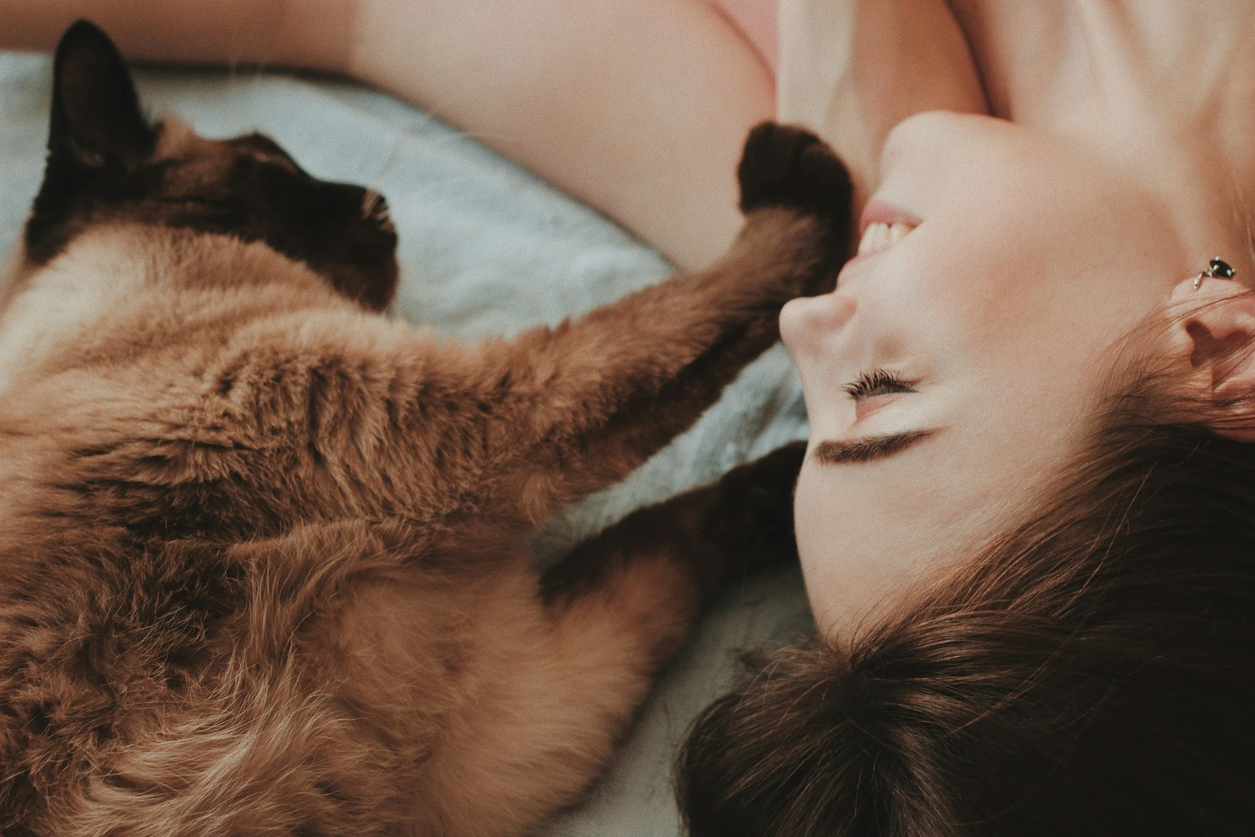 A person with long brown hair and earrings lying on a bed, smiling with closed eyes, next to a brown and black cat laying on its back with one paw stretched out towards the person.