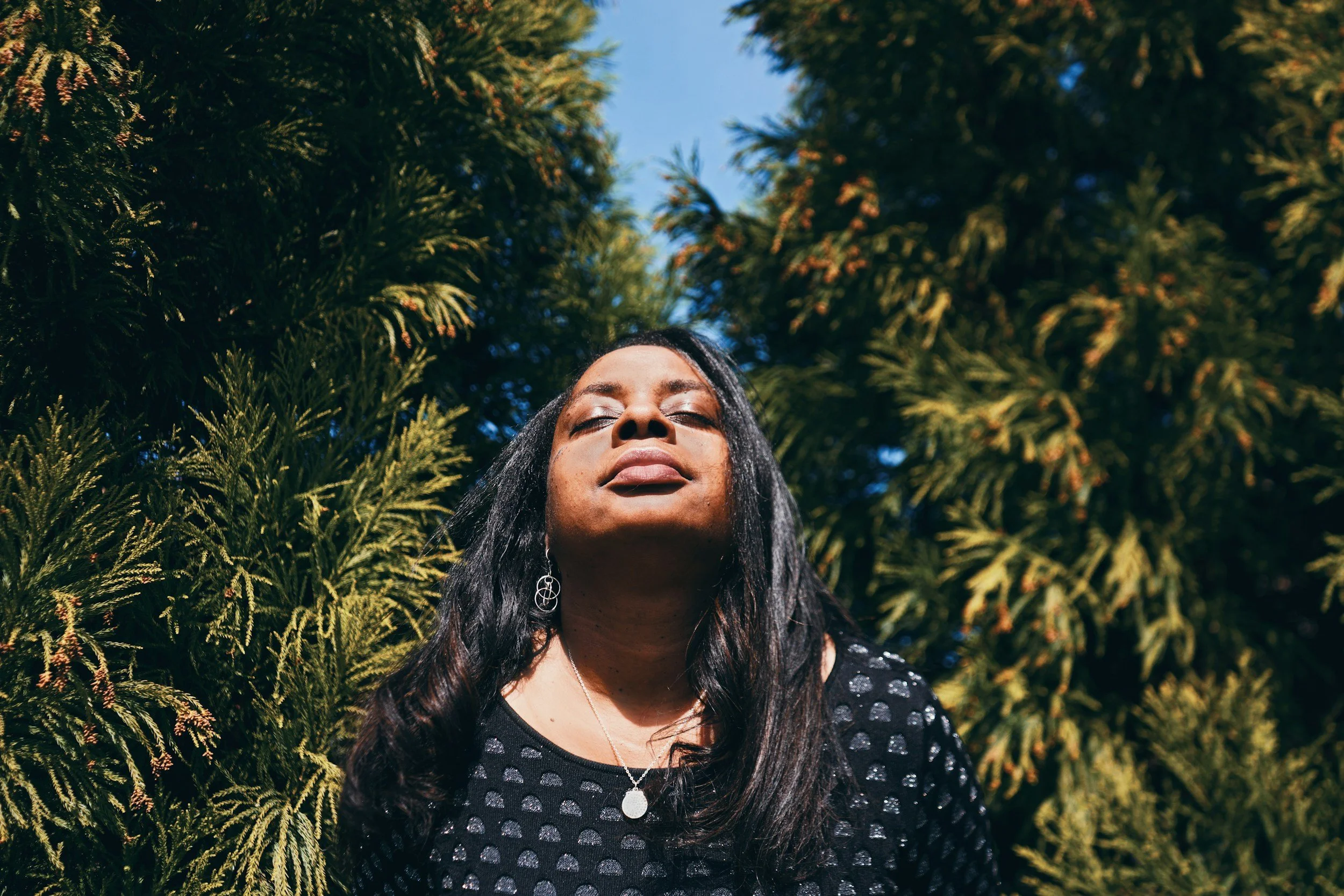 A woman with long dark hair and earrings stands outdoors with her eyes closed, surrounded by green trees against a blue sky.