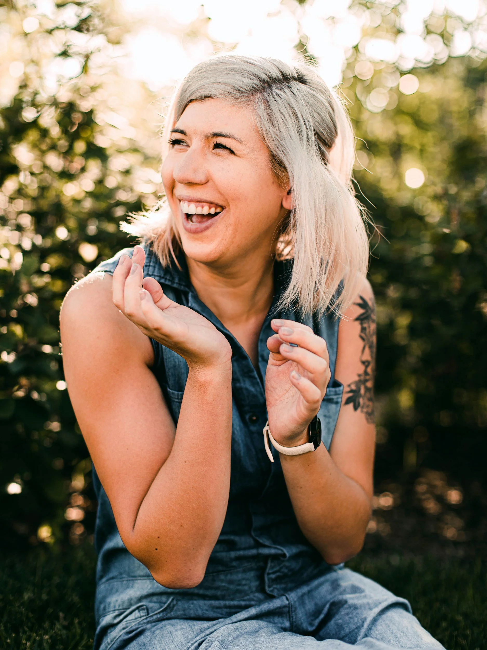 A smiling woman with short gray hair and tattoos, wearing a sleeveless denim shirt, sitting outdoors in a park or garden during sunset, with sunlight filtering through the trees in the background.