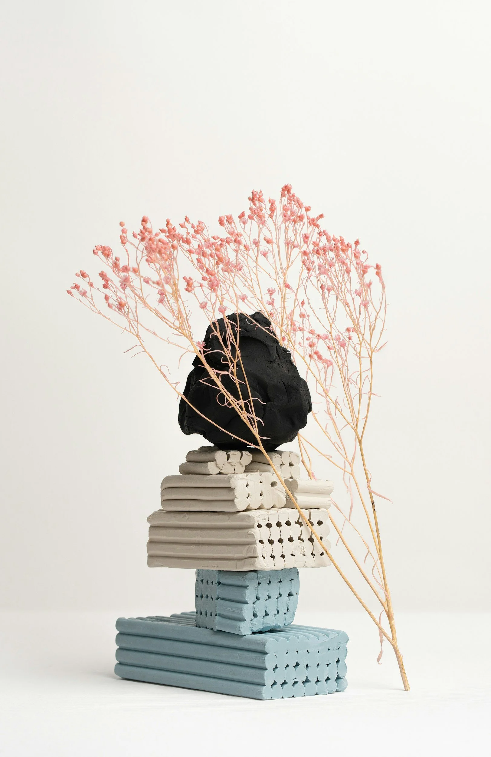 Modern abstract sculpture made of stacked geometric shapes in neutral and pastel colors, topped with a black textured element, adorned with pink dried flowers, set against a plain white background.