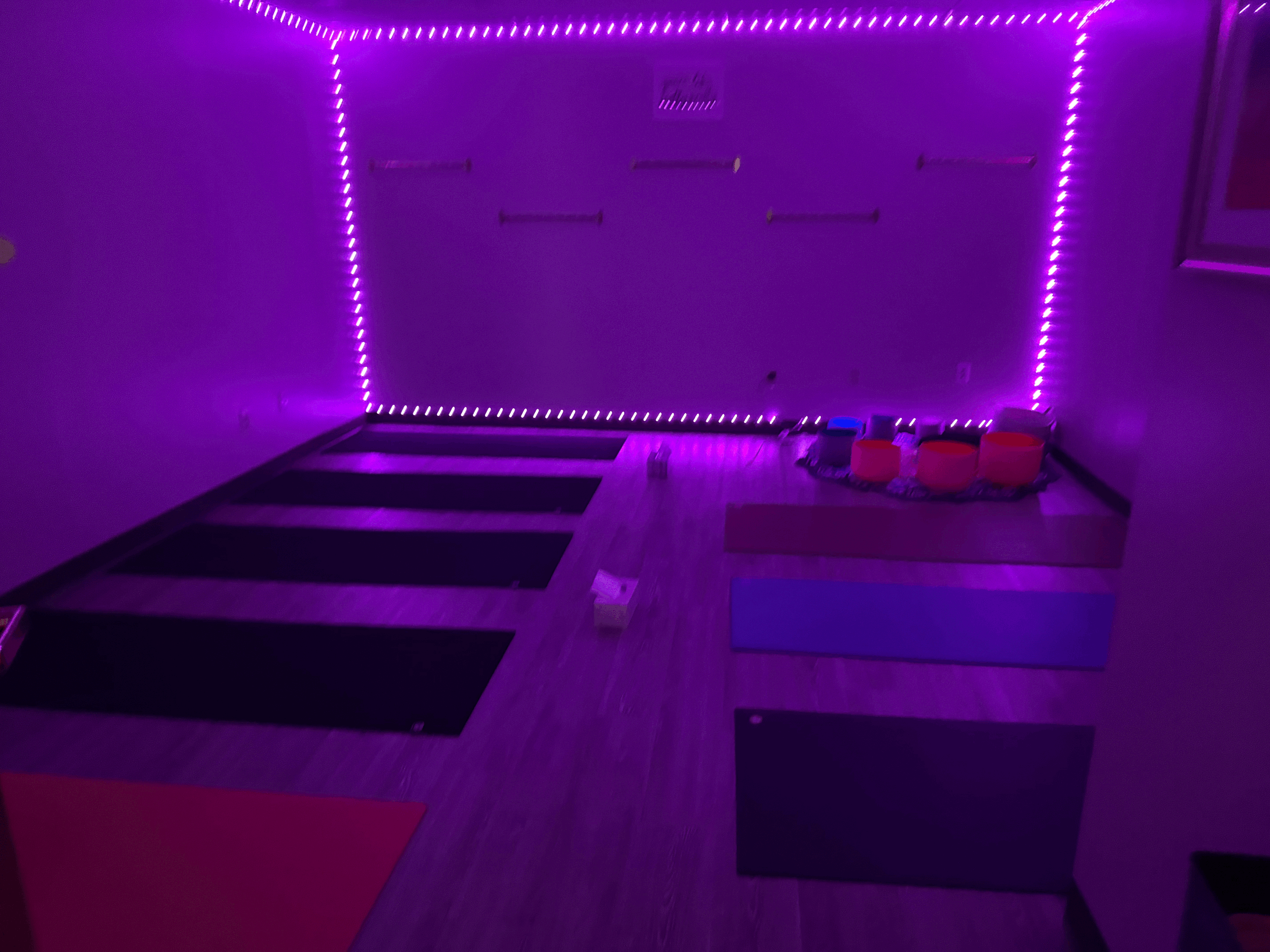 A yoga or exercise room with purple LED strip lighting around the ceiling edges, black yoga mats on the wooden floor, and some smaller mats and exercise props on a foam block in the corner.