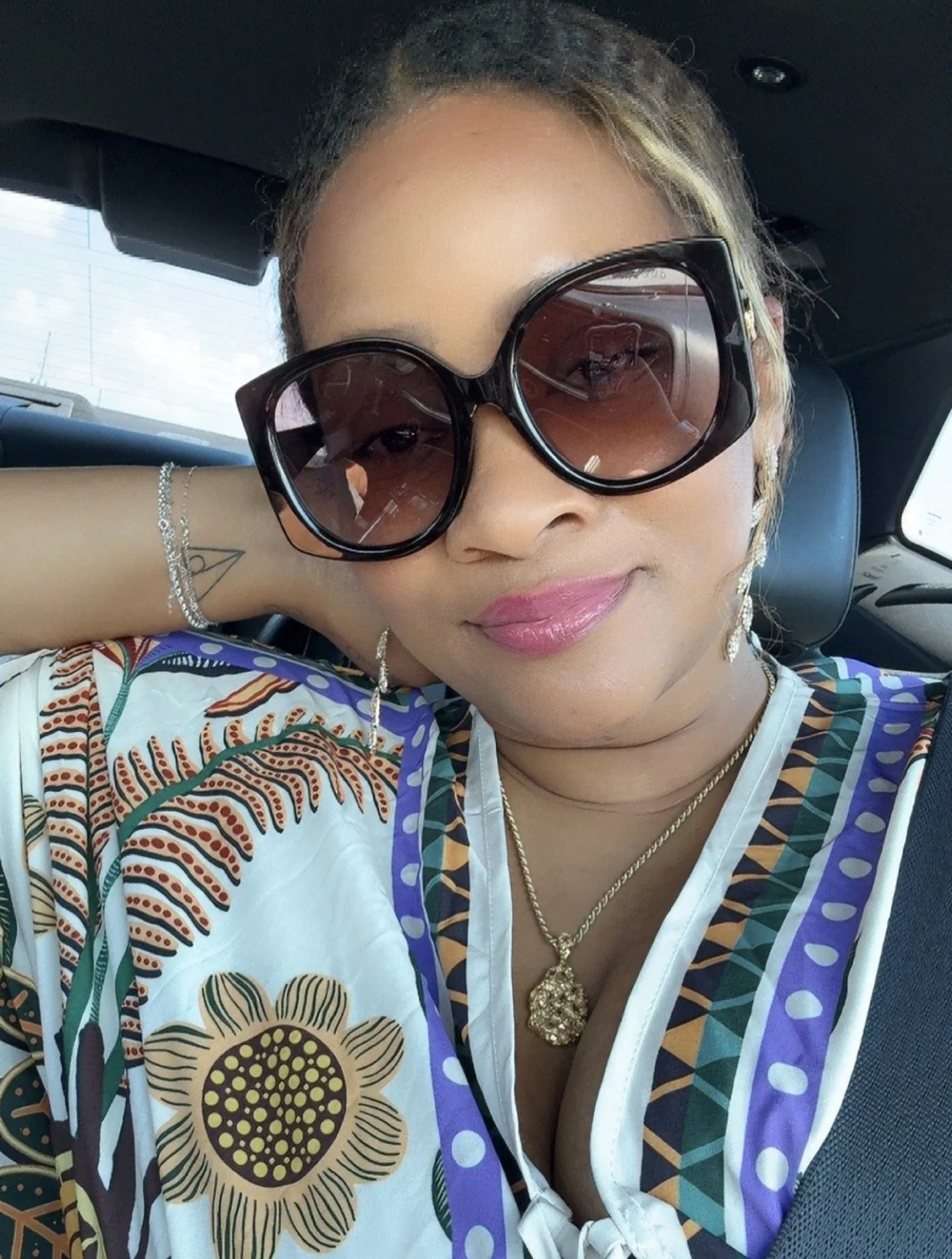 A woman wearing large sunglasses, earrings, a colorful patterned blouse, a gold necklace, and a bracelet, sitting in a car.