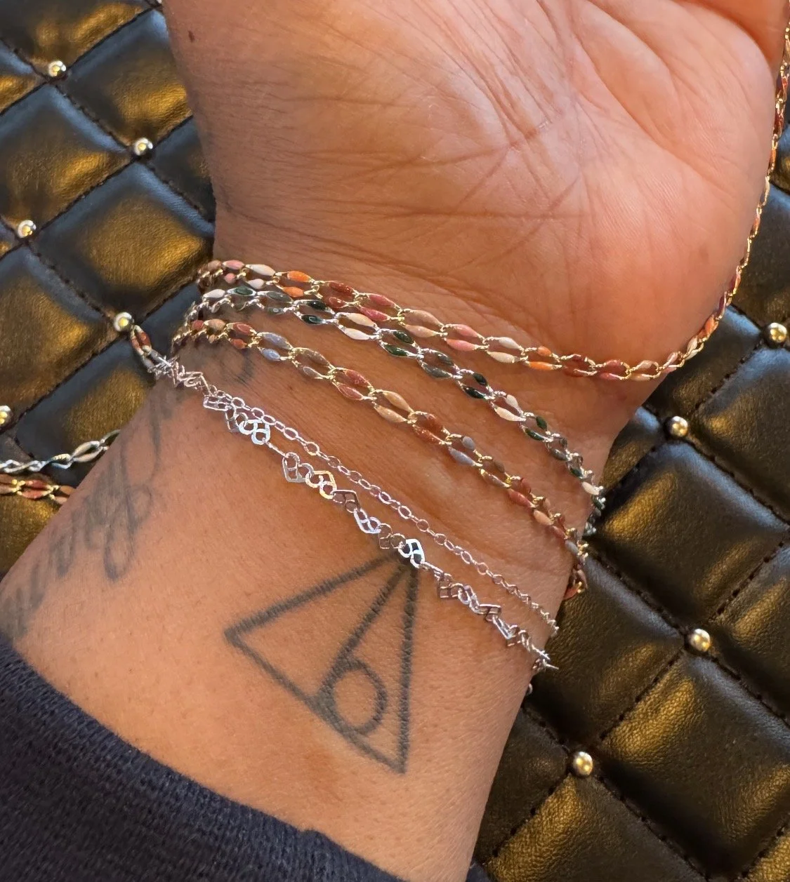 A close-up of a person's wrist wearing multiple delicate chain bracelets with colorful and silver links, and a tattoo of a pyramid with an eye inside, with a black quilted leather surface with gold accents in the background.