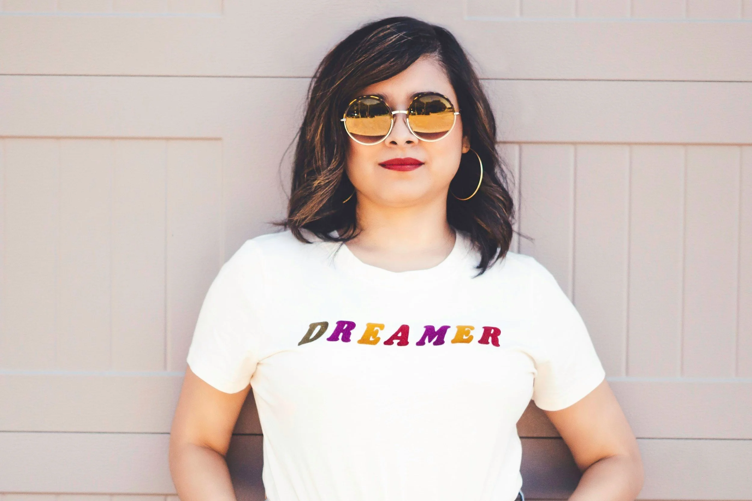 Woman with dark wavy hair in sunglasses, a white T-shirt with the word "DREAMER" in colorful letters, standing against a beige wall.