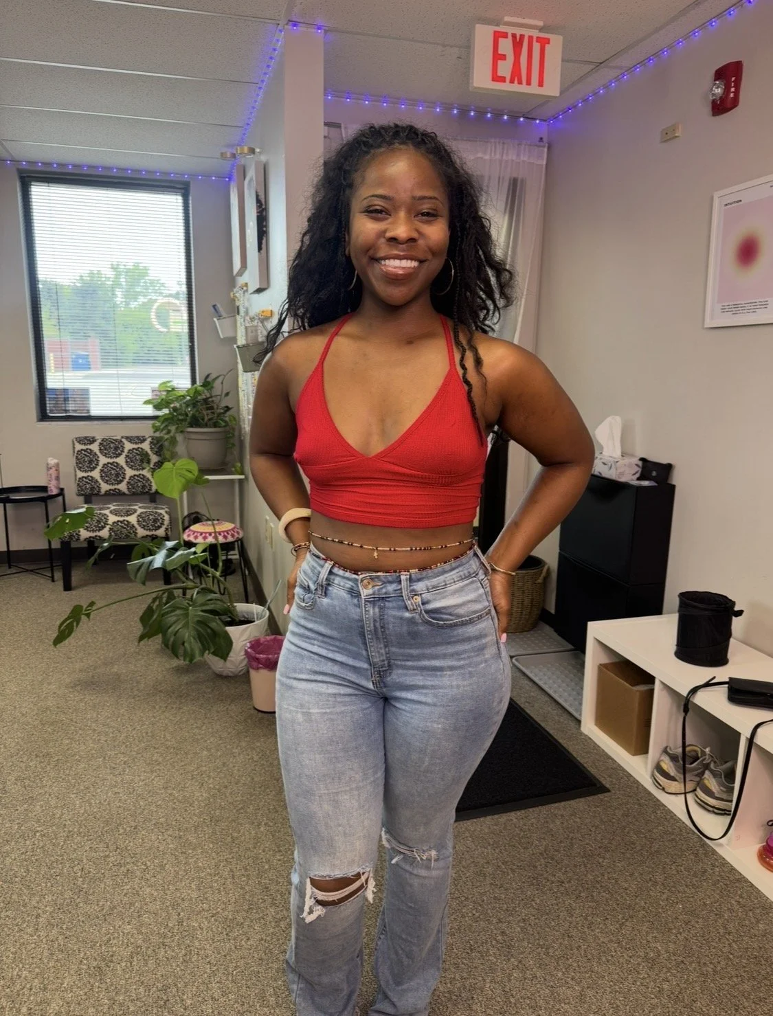A woman with curly hair wearing a red crop top and ripped jeans, standing indoors near potted plants wearing new waistbeads. Create your own waistbeads for girls night out, private parties and DIY waist beads.