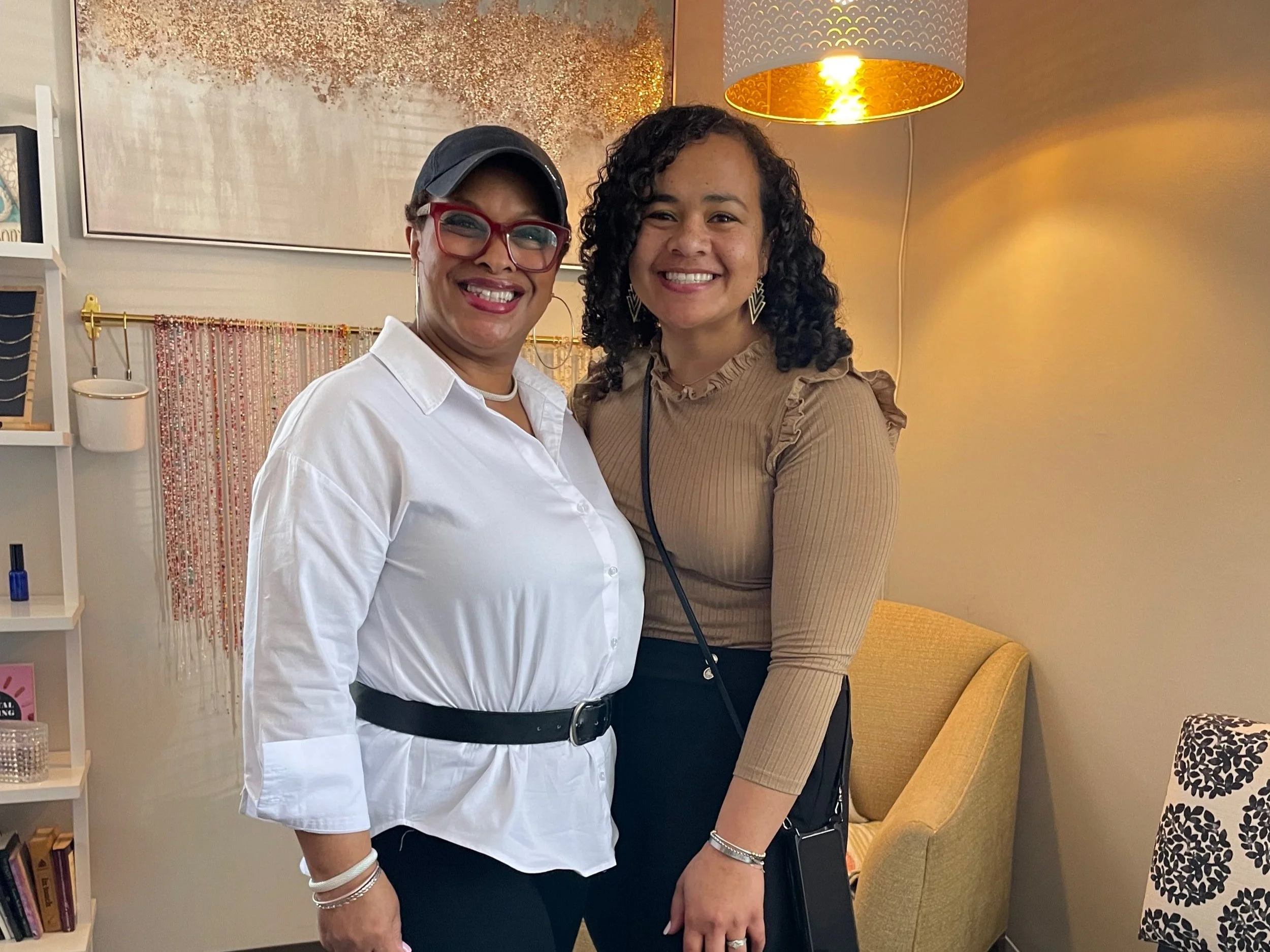 Two women standing close together, smiling for the camera. One woman is wearing a white button-up shirt, glasses, and a baseball cap, while the other is wearing a beige long-sleeve top with ruffle details and a black skirt. They are in a well-decorated indoor setting with a yellow armchair and various decorative items in the background.