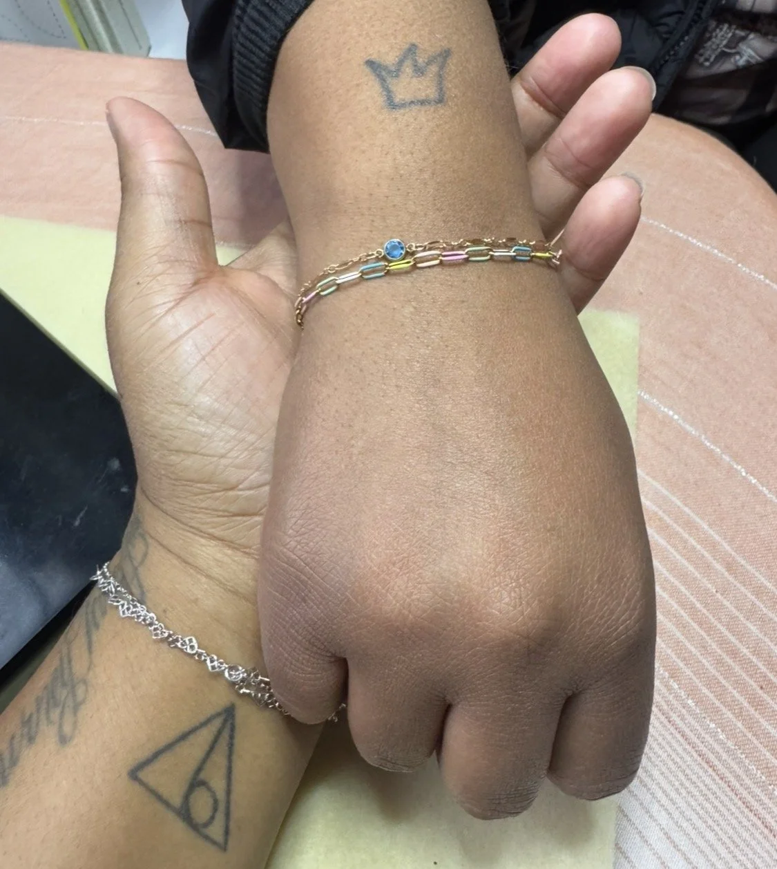A person's hand resting on their own wrist, wearing a silver bracelet with intricate design. The person's arm features a small tattoo and a delicate chain bracelet. The other person's hand, with a colorful bracelet and a small blue gem, is gently hol
