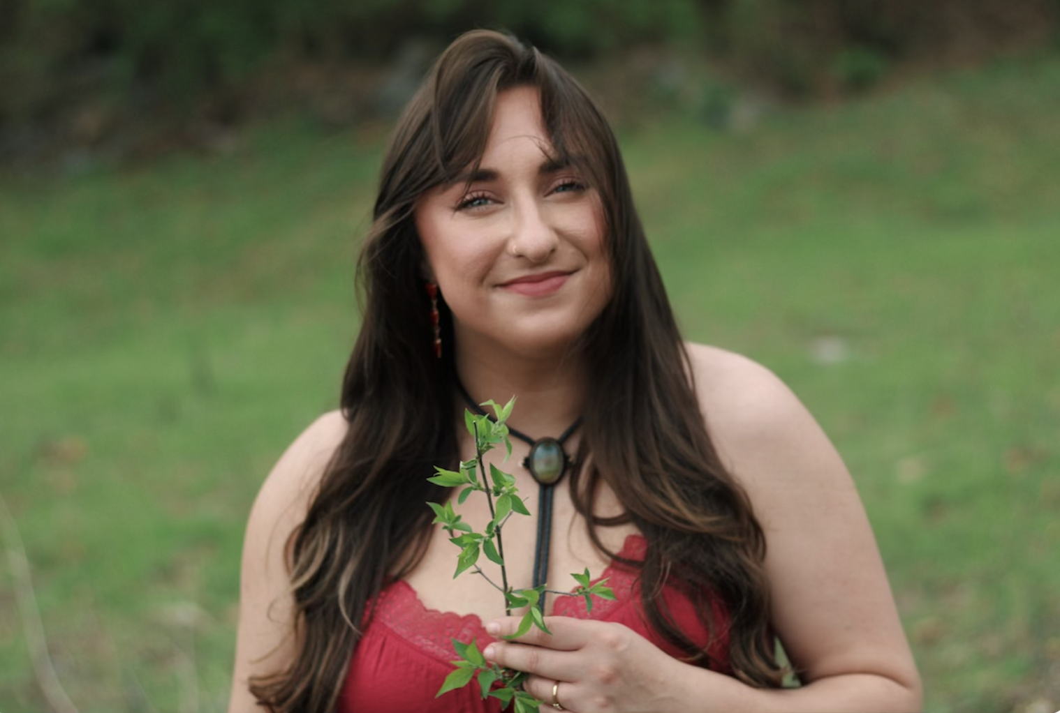 A woman with long dark hair, wearing a red dress, holding a green leafy branch, standing outdoors with a blurred green background