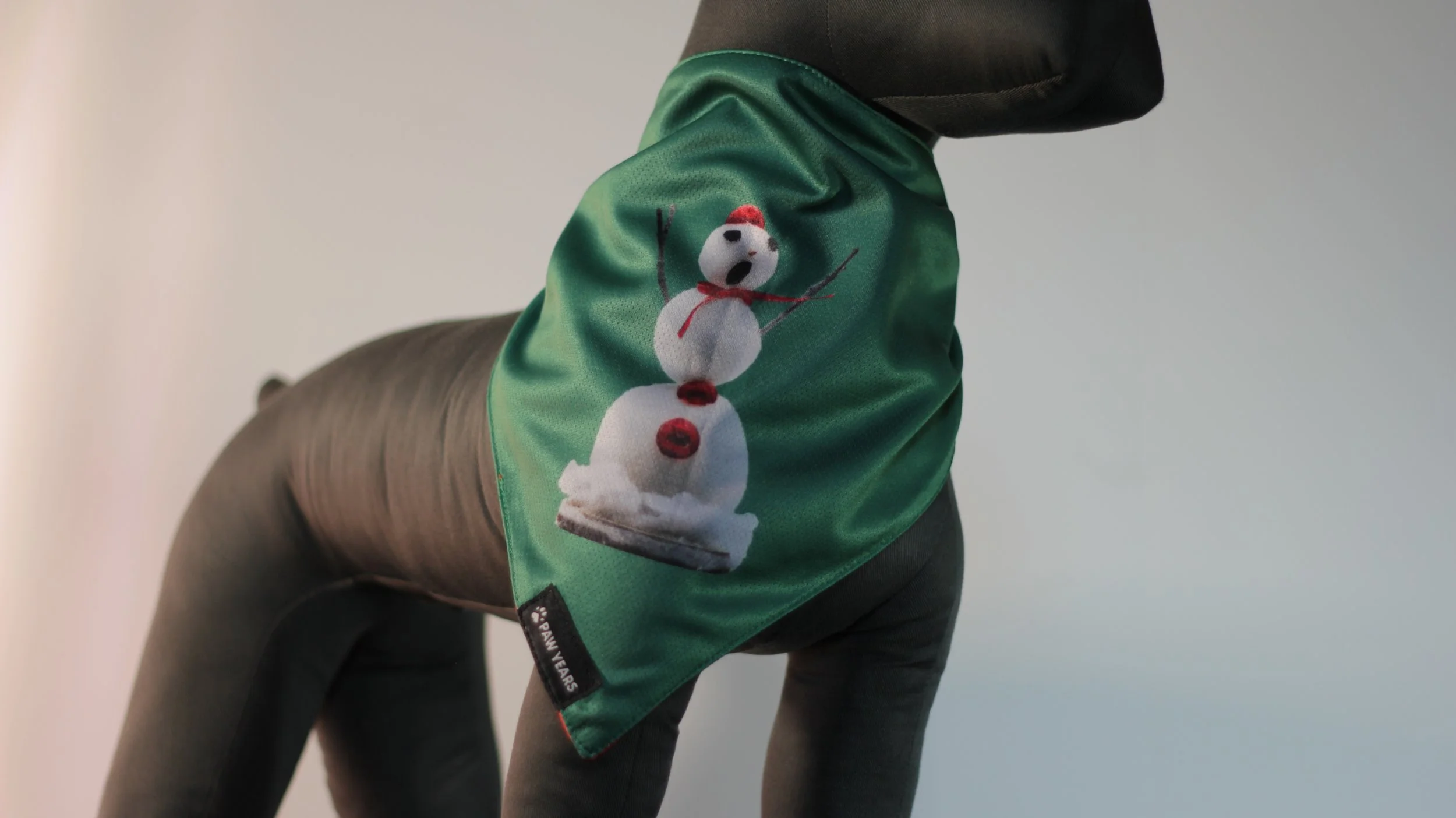 Small dog mannequin wearing the green side of the Winter Love limited edition bandana with snowman design.