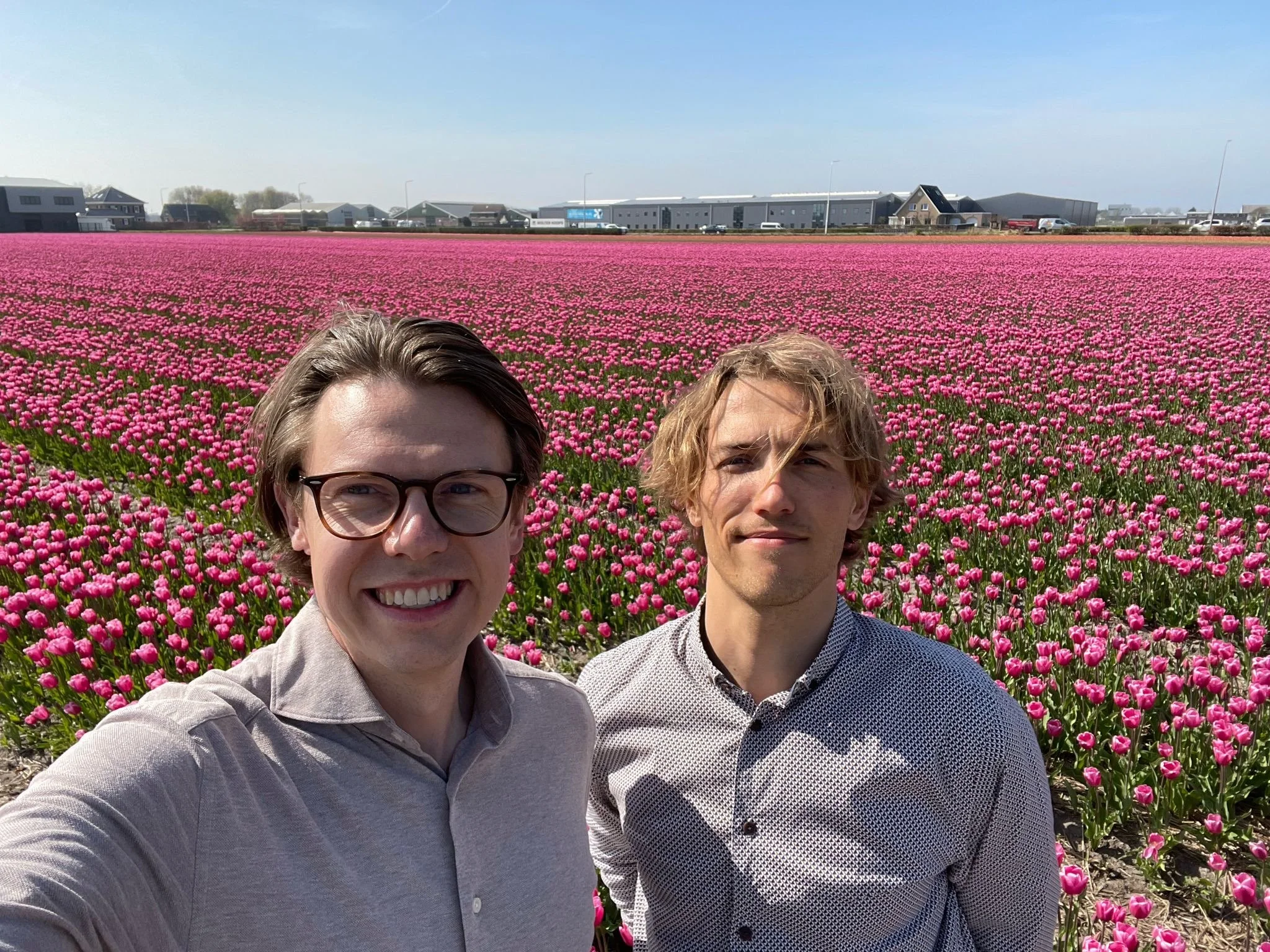 Two young men taking a selfie in front of a vast field of pink tulips under a clear blue sky.