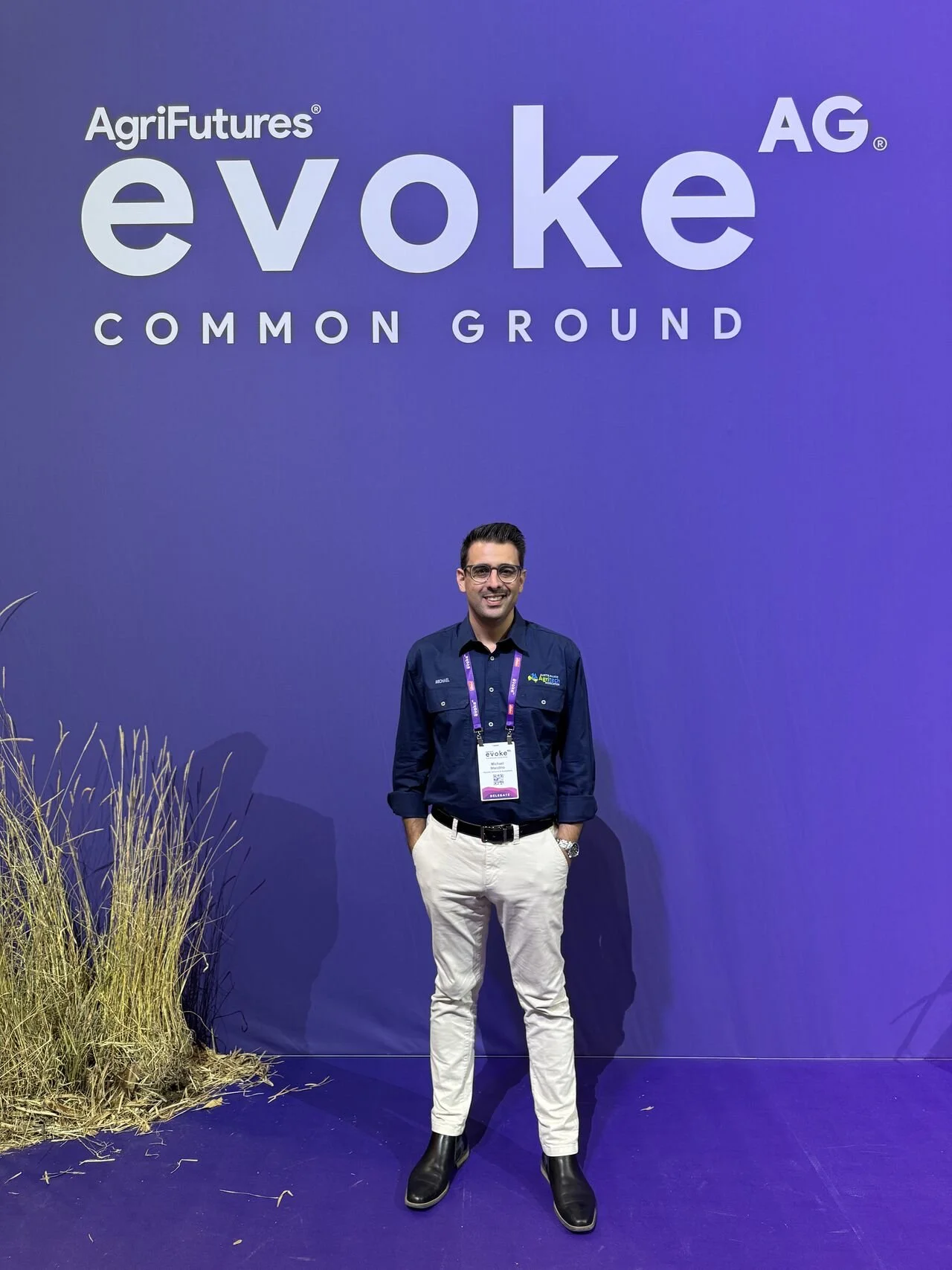 A man standing in front of a purple wall with the words 'AgriFutures evoke COMMON GROUND'. He is wearing glasses, a dark blue shirt, beige pants, and black boots.